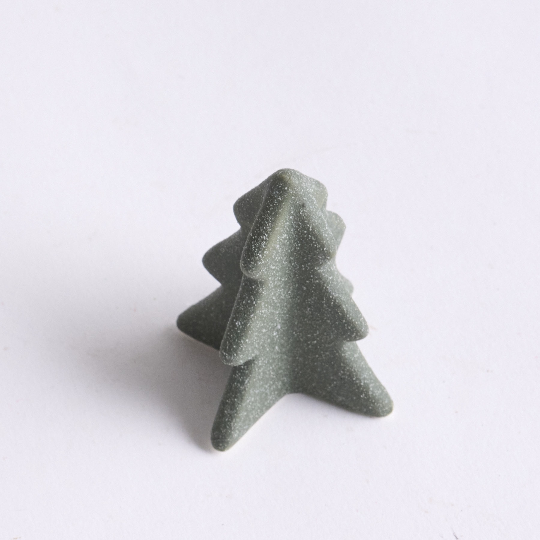 Auf einem schlichten weißen Hintergrund befindet sich ein kleines, stilisiertes baumförmiges Objekt aus graugrünem Material.