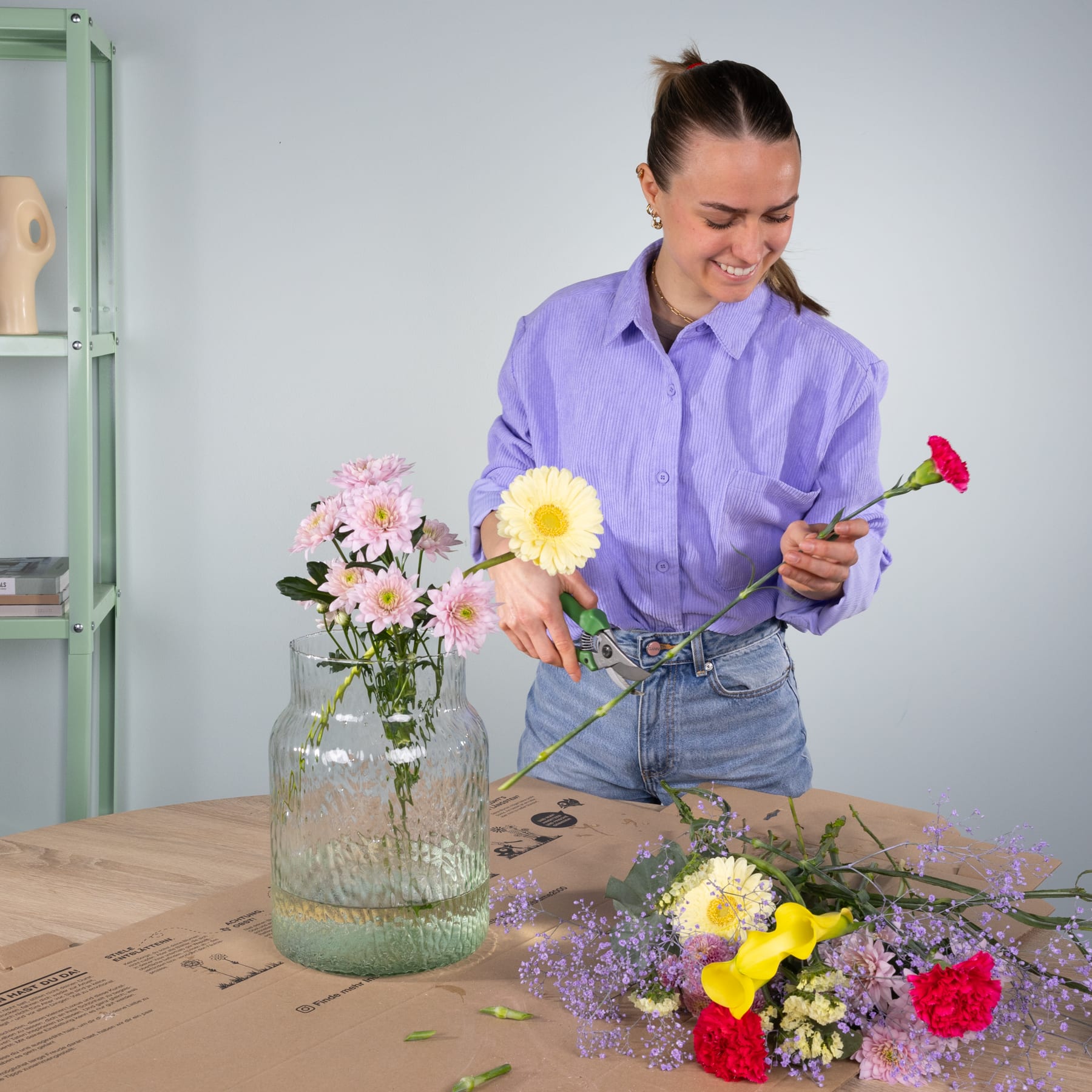 Eine junge Frau in einem lila Hemd arrangiert einen Strauß bunter Blumen auf einem Holztisch in einem Raum mit einem grünen Regal im Hintergrund.