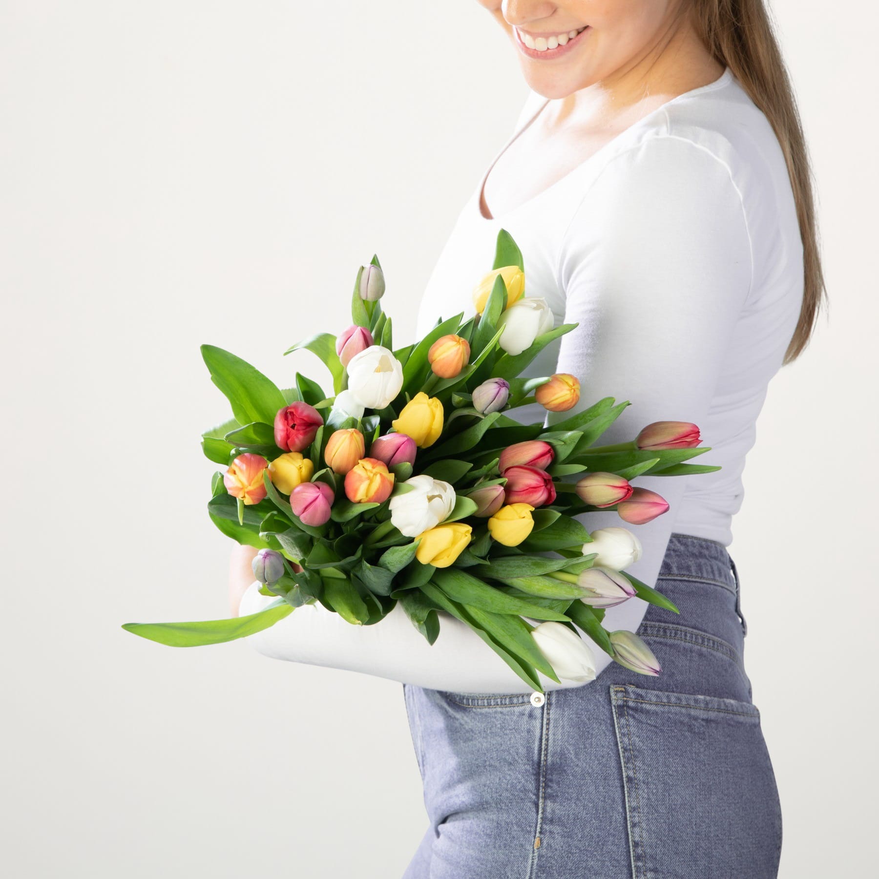 Eine junge Frau in weißem Hemd und Jeans hält einen leuchtenden Strauß bunter Tulpen vor einem schlichten weißen Hintergrund.