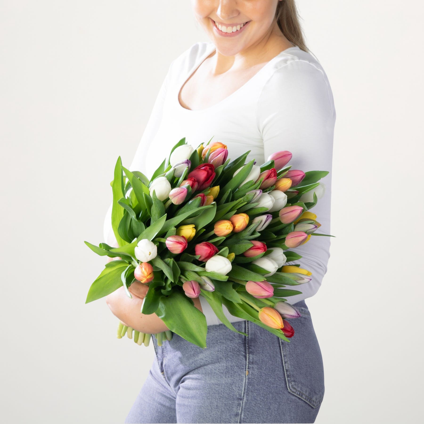 Eine lächelnde Frau in einem weißen Oberteil hält einen leuchtenden Strauß bunter Tulpen vor einem schlichten weißen Hintergrund.