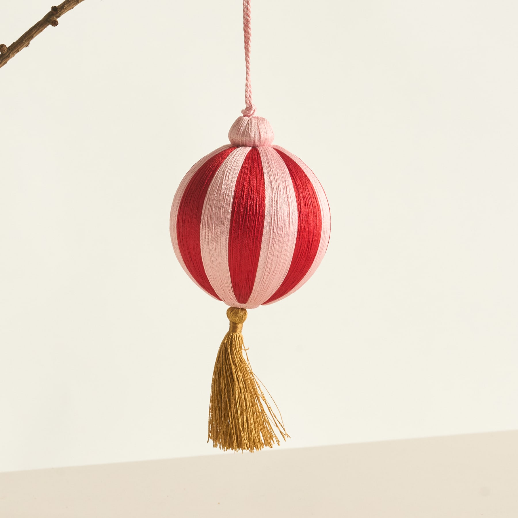 Ein leuchtend rot-weiß gestreiftes Ornament mit einer goldenen Quaste hängt vor einem schlichten, hellen Hintergrund an einem kahlen Ast.