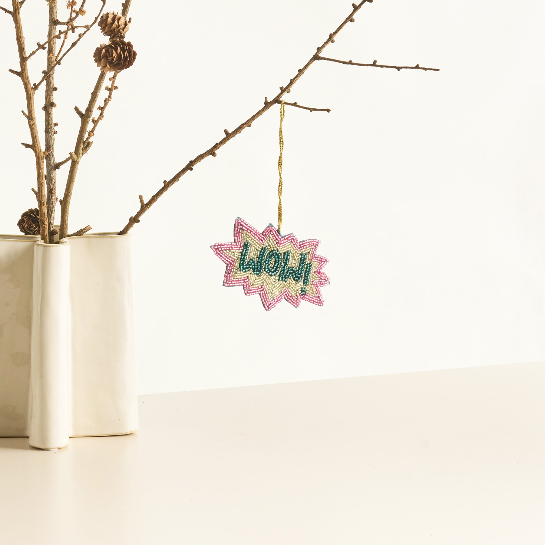 In einer weißen Keramikvase hängt vor einem schlichten weißen Hintergrund ein Zweig mit einem dekorativen „Wow!“-Ornament daran.