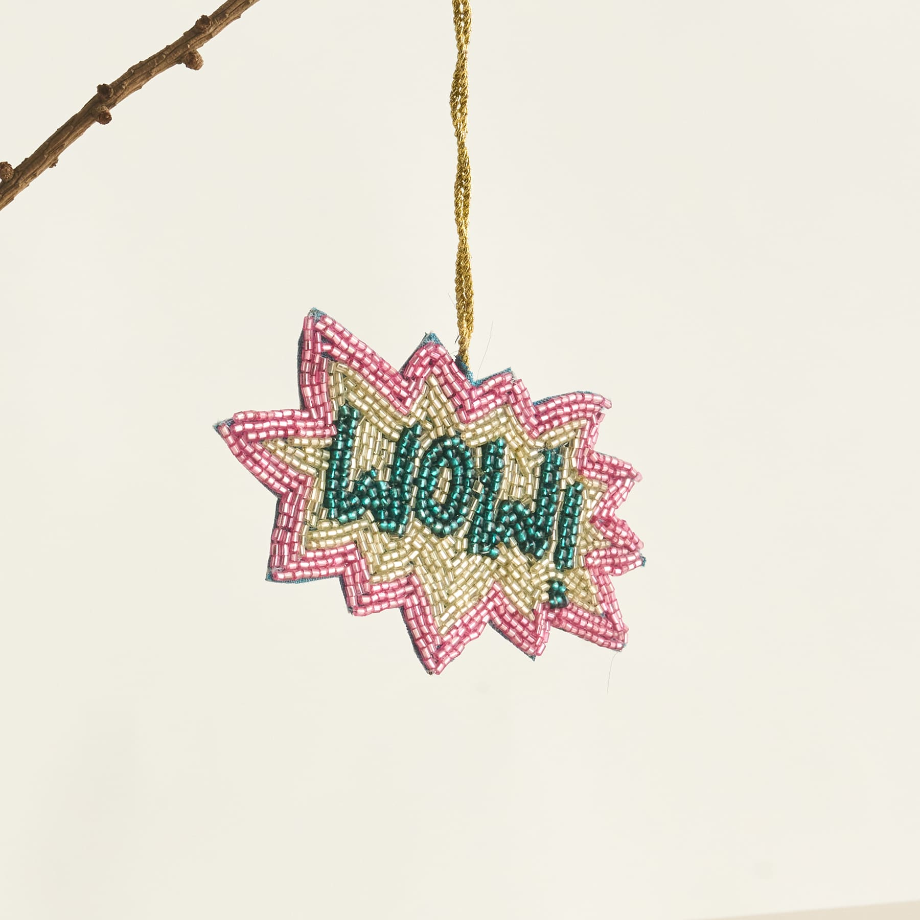 Ein buntes, mit Perlen besetztes Ornament in Form eines Sterns oder einer Explosion, in dessen Mitte das Wort „Wow!“ steht, hängt vor einem schlichten, hellen Hintergrund an einem Holzast.