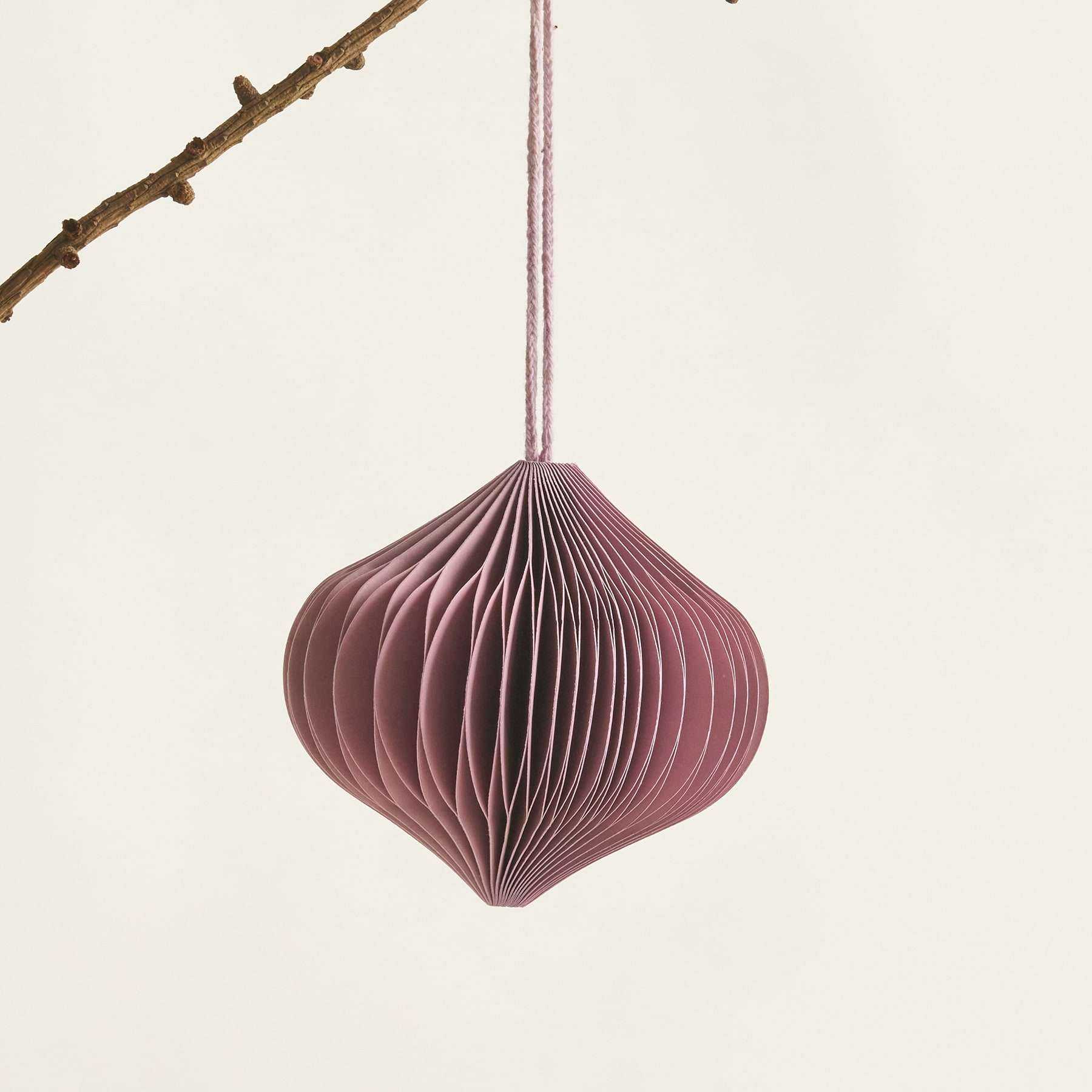 Ein zartes, gefaltetes Papierornament in einem sanften Rosaton hängt vor einem schlichten, cremefarbenen Hintergrund an einem kahlen Ast.