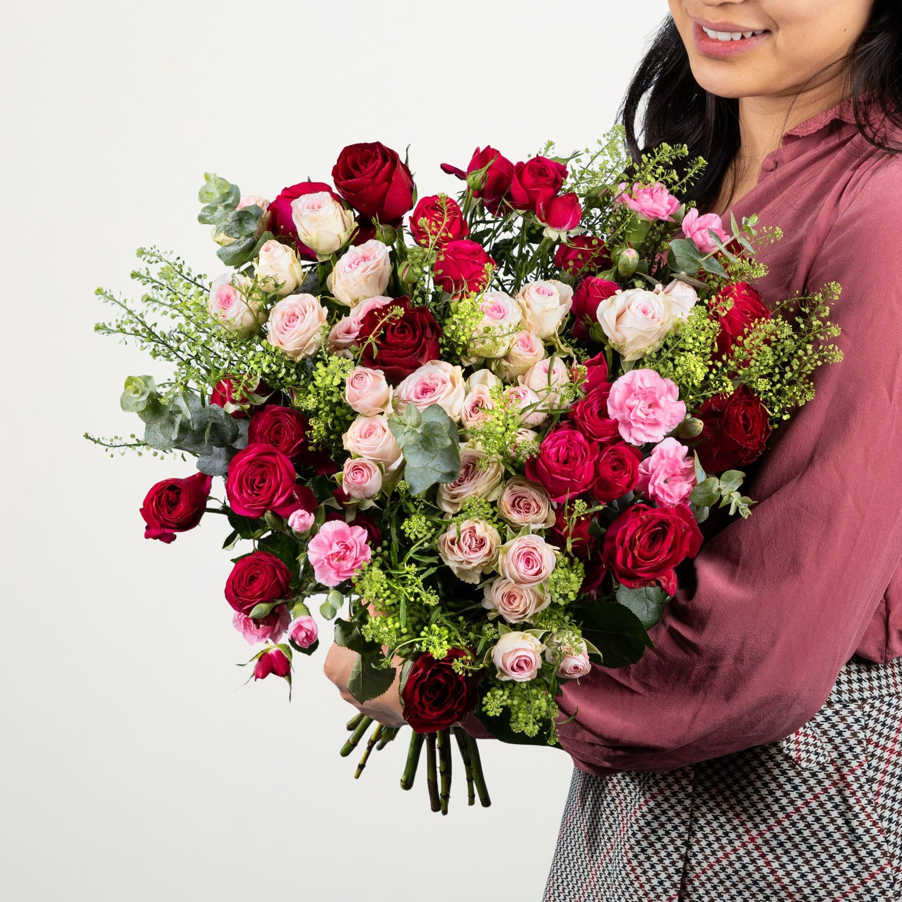 Ein großer, leuchtender Strauß roter Rosen und rosa Nelken, gehalten von einer Person in einem burgunderfarbenen Hemd vor einem schlichten weißen Hintergrund.
