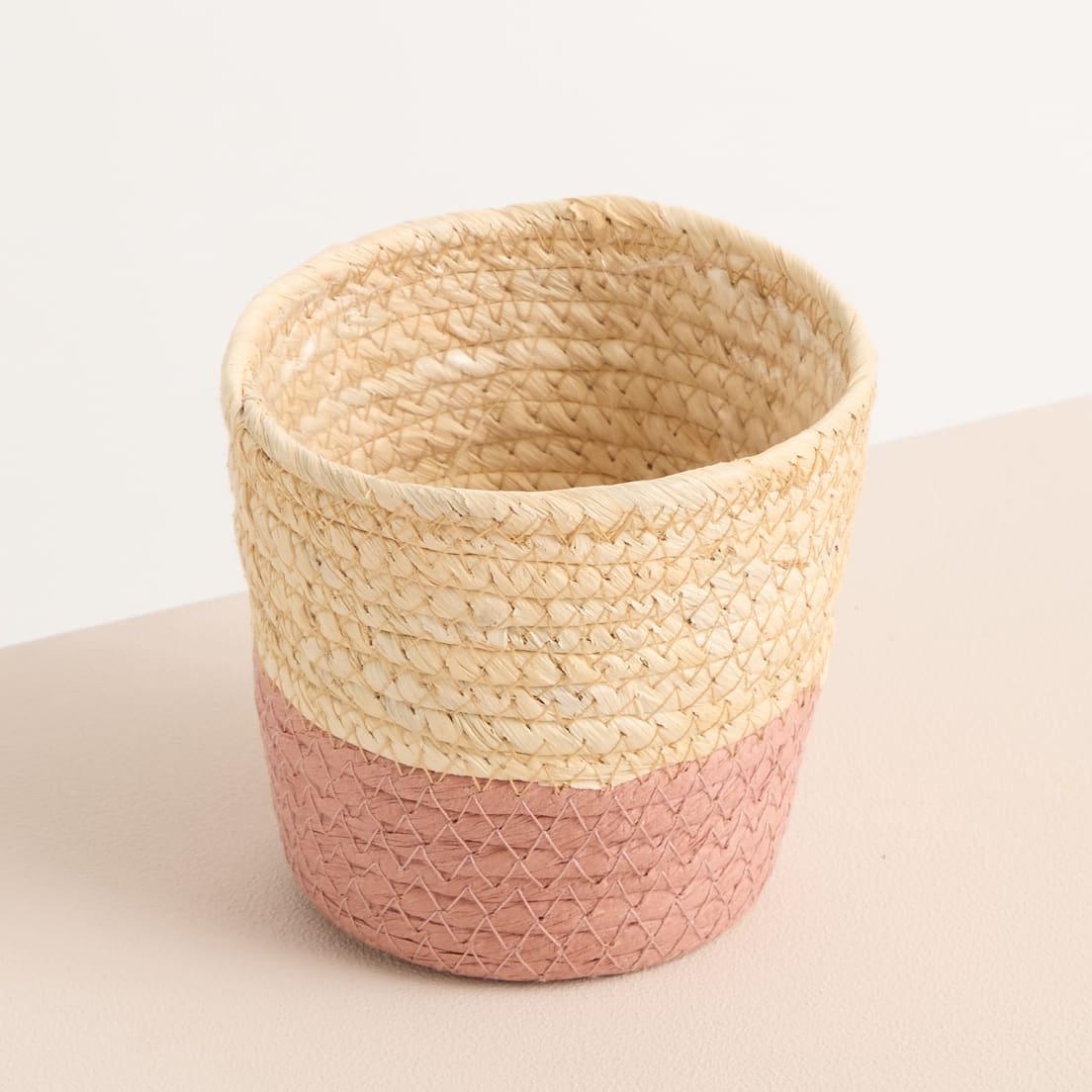 Ein geflochtener Korb in natürlichem Beige mit einem rosa geometrischen Muster am Boden, der auf einer hellen Oberfläche steht.