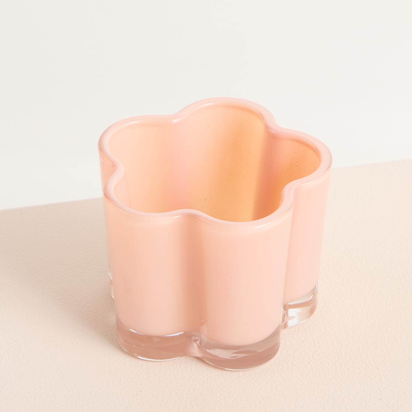 Eine zarte, pfirsichfarbene Glasvase mit Blumenform steht auf einer schlichten, hellen Oberfläche.