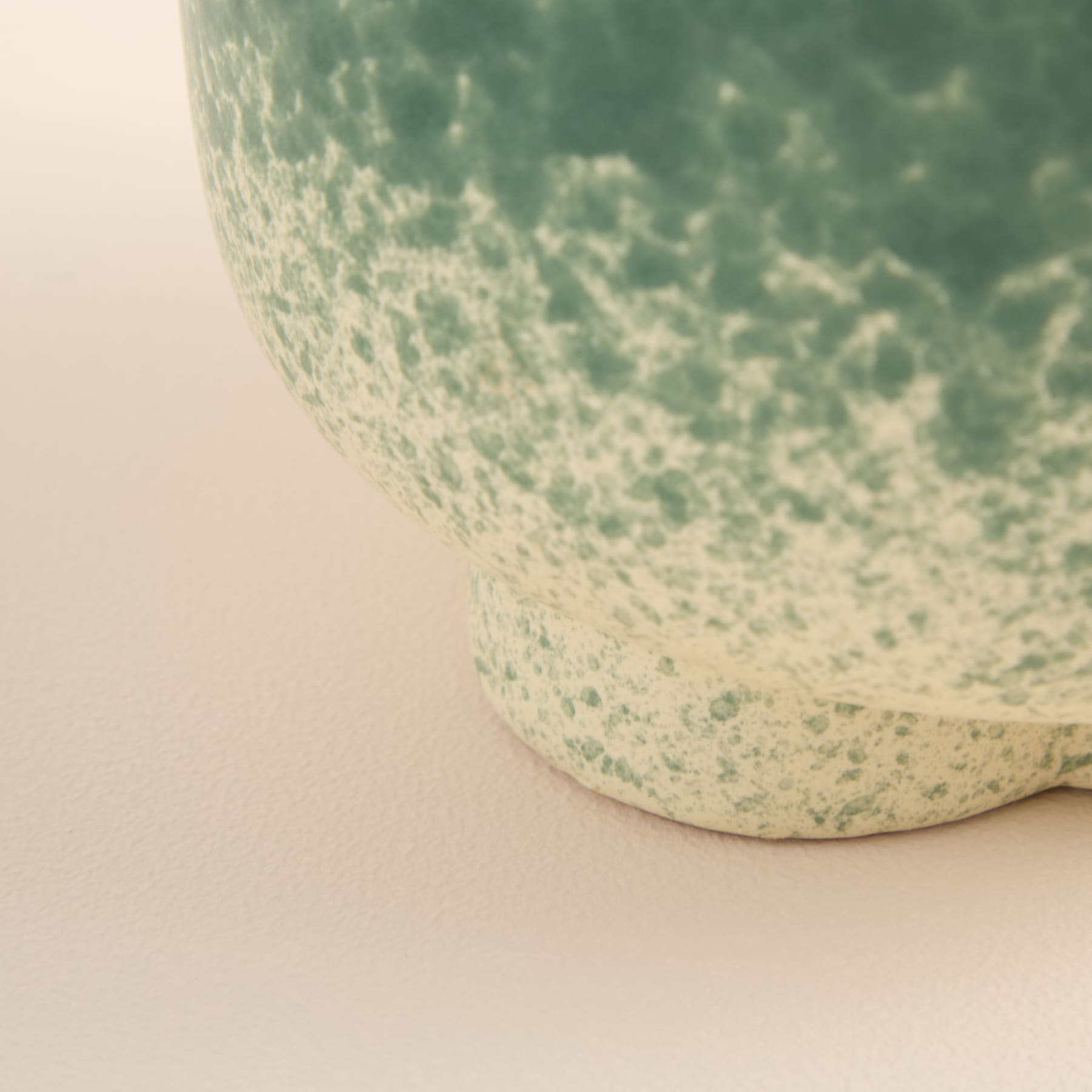 Eine Nahaufnahme einer Keramikschale oder eines Keramikgefäßes mit einer gesprenkelten grün-weißen Glasur, das auf einer hellen Oberfläche steht.