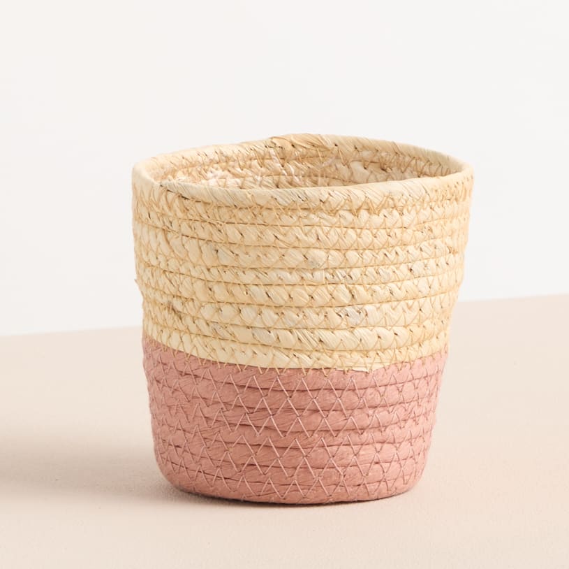 Ein geflochtener Korb mit einer natürlichen beigen Farbe an der Oberseite und einem rosa geometrischen Muster an der Unterseite, der auf einer hellen Oberfläche steht.