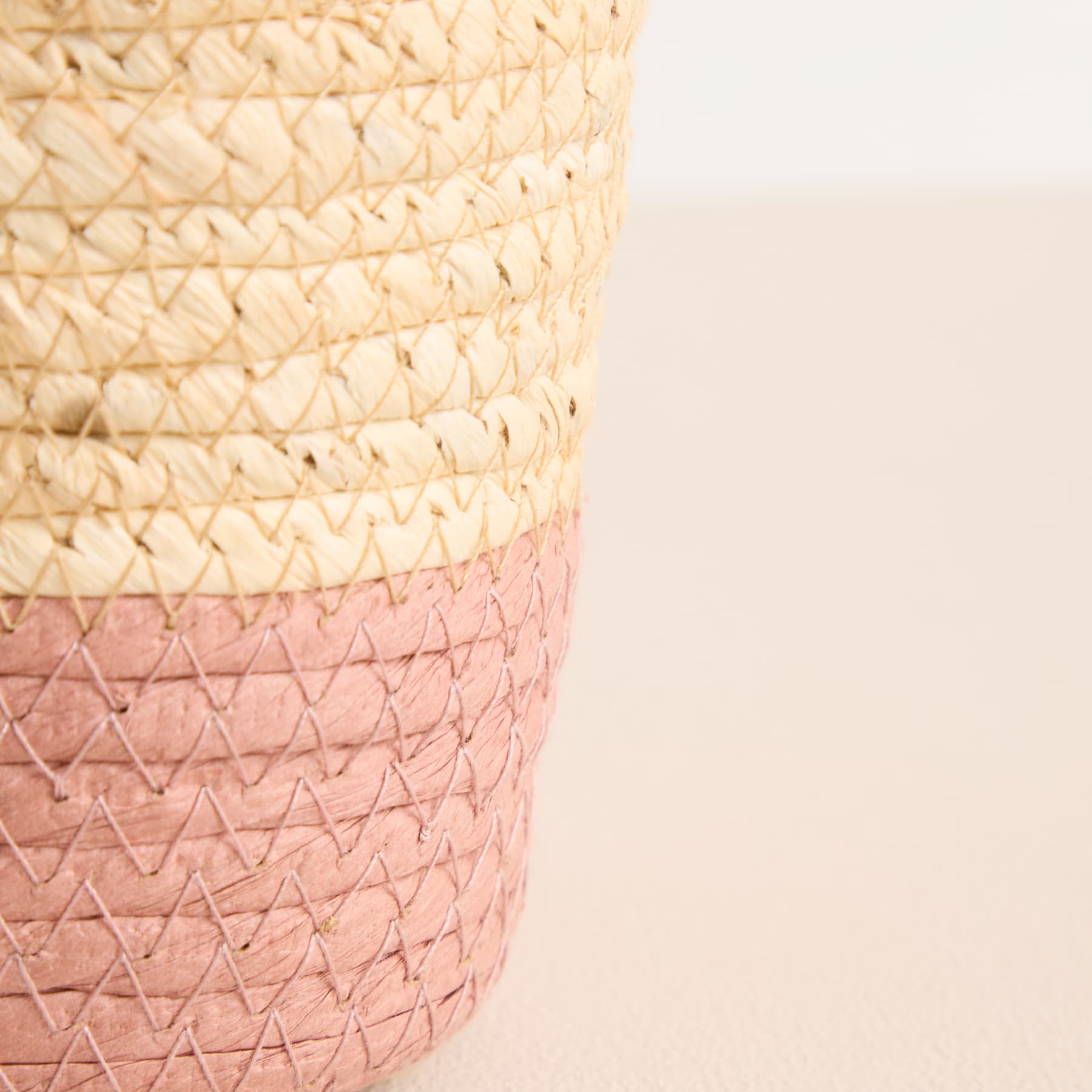Ein geflochtener Korb oder Behälter mit einem Farbverlauf, der von einem hellen Beige oben zu einem zarten Rosa unten übergeht, vor einem schlichten, hellen Hintergrund.