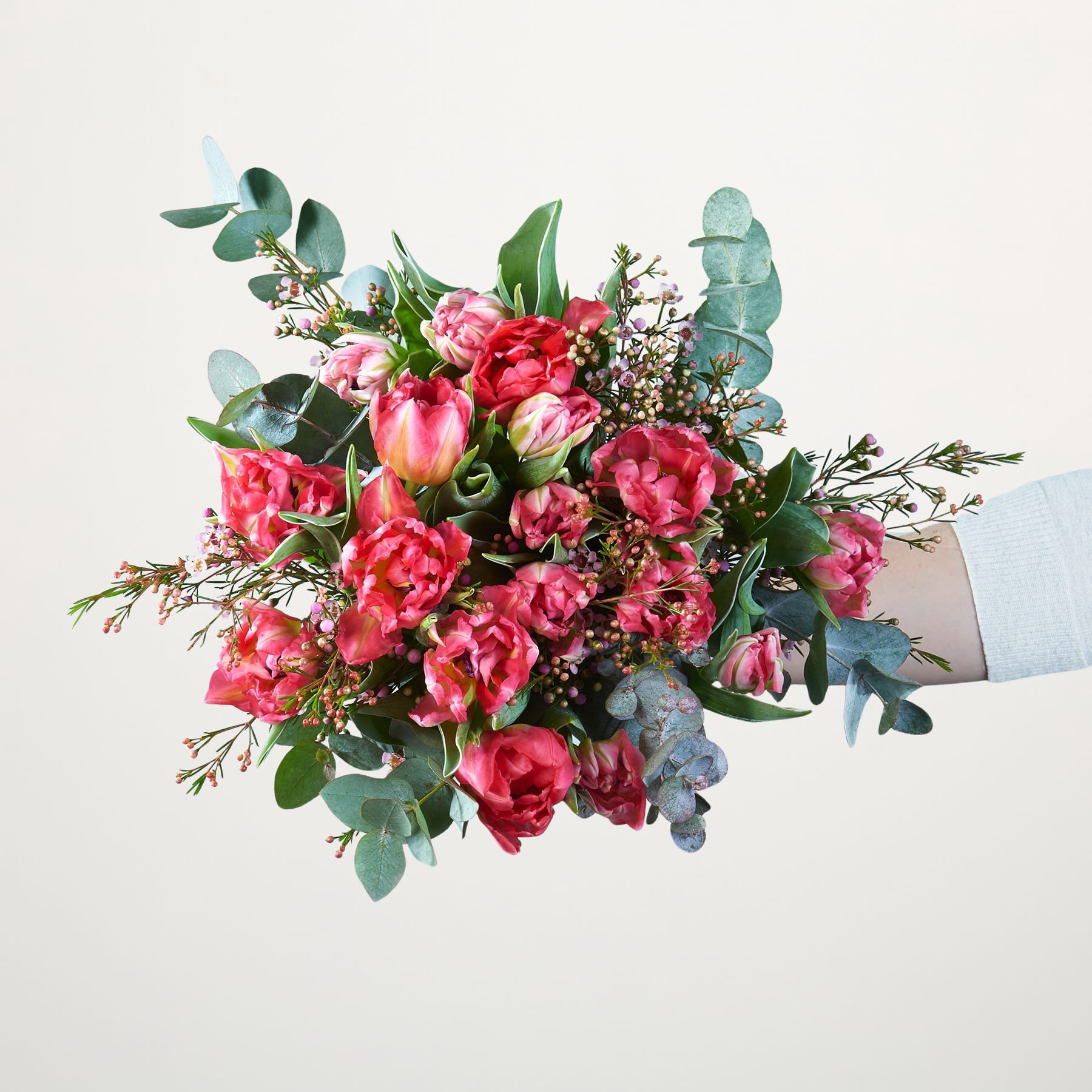 Ein leuchtender Strauß üppiger, rosa Pfingstrosen und anderer bunter Blumen, akzentuiert mit Grün, wird vor einem schlichten weißen Hintergrund in den Händen einer Person gehalten.