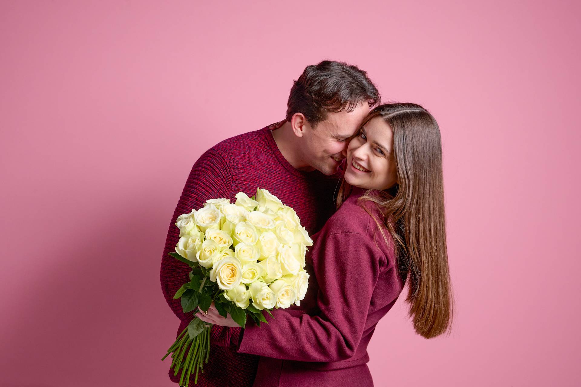Ein junges Paar umarmt sich und lächelt, wobei die Frau einen Strauß weißer Blumen vor einem leuchtend rosa Hintergrund hält.