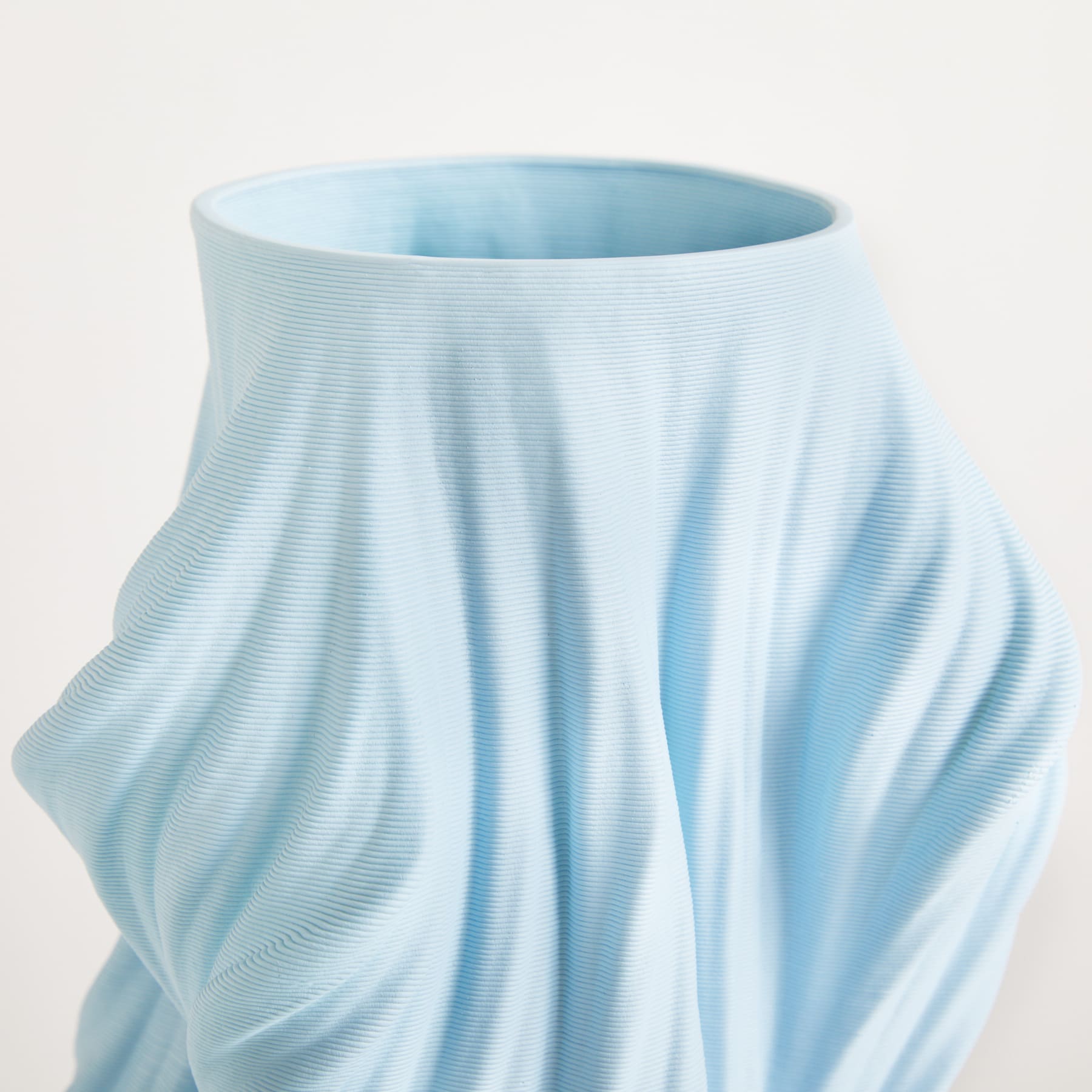 Eine hellblaue, plissierte Keramikvase mit einer glatten, fließenden Textur vor einem schlichten weißen Hintergrund.