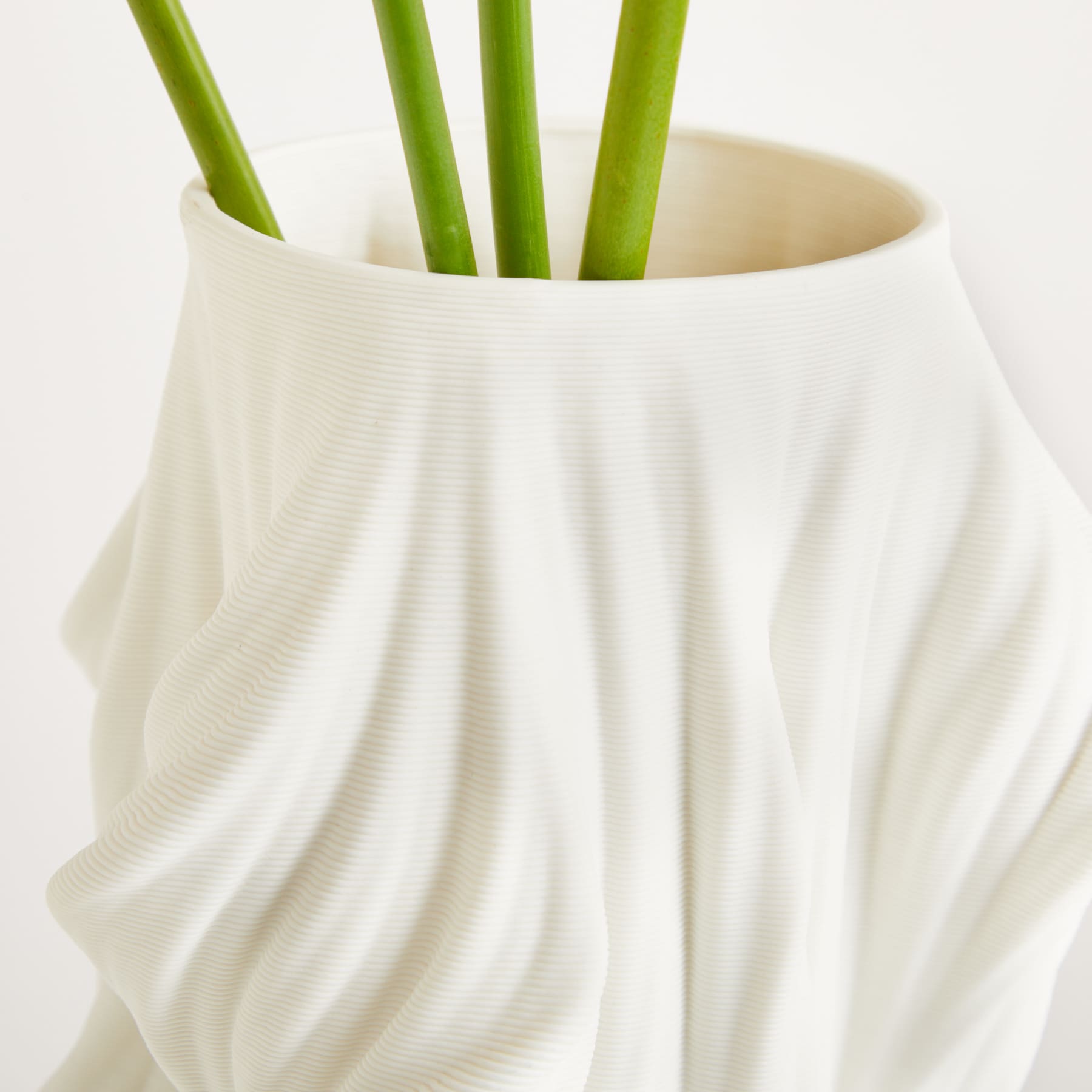 Eine weiße Keramikvase oder ein weißer Keramikbehälter enthält mehrere grüne, schlanke Stiele, möglicherweise Aloe Vera oder eine ähnliche Pflanze, vor einem schlichten, hellen Hintergrund.