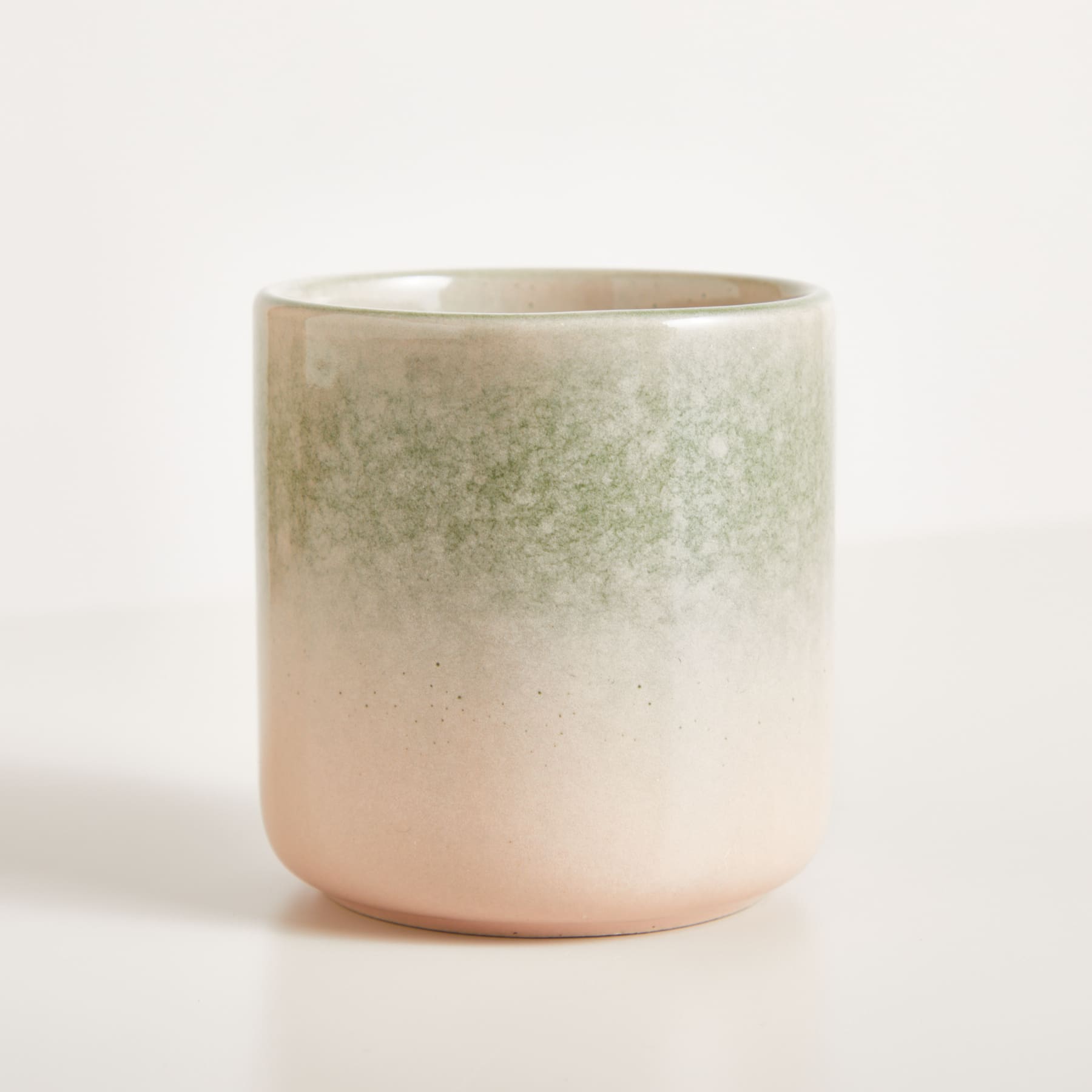 Auf einer schlichten, hellen Oberfläche steht eine Keramiktasse oder ein Keramikbecher mit einem Farbverlauf aus hellen und dunklen Farben, von blassem Rosa bis zu gedämpftem Grün.