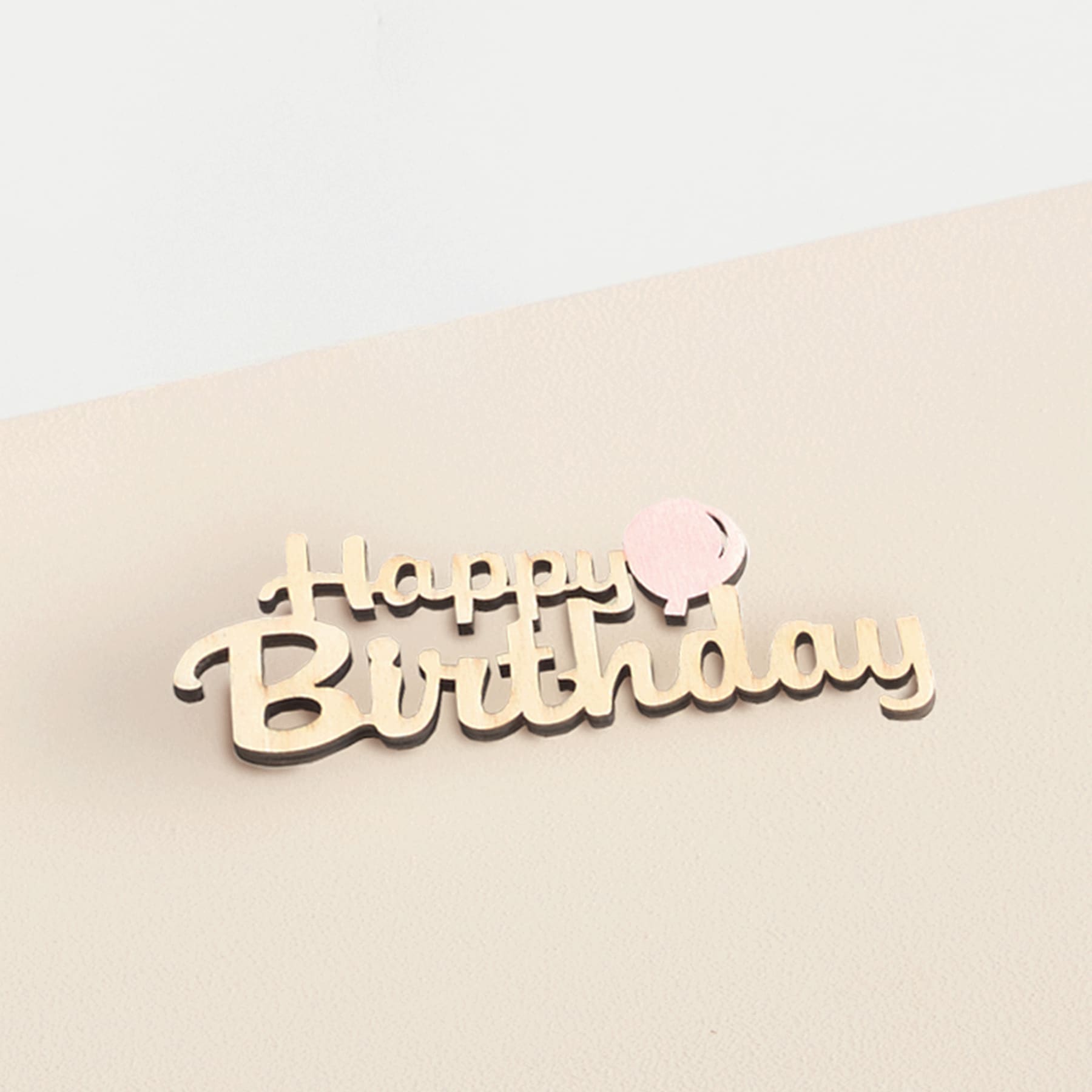 Das Bild zeigt ein hölzernes „Happy Birthday“-Schild vor einem hellbeigen Hintergrund.