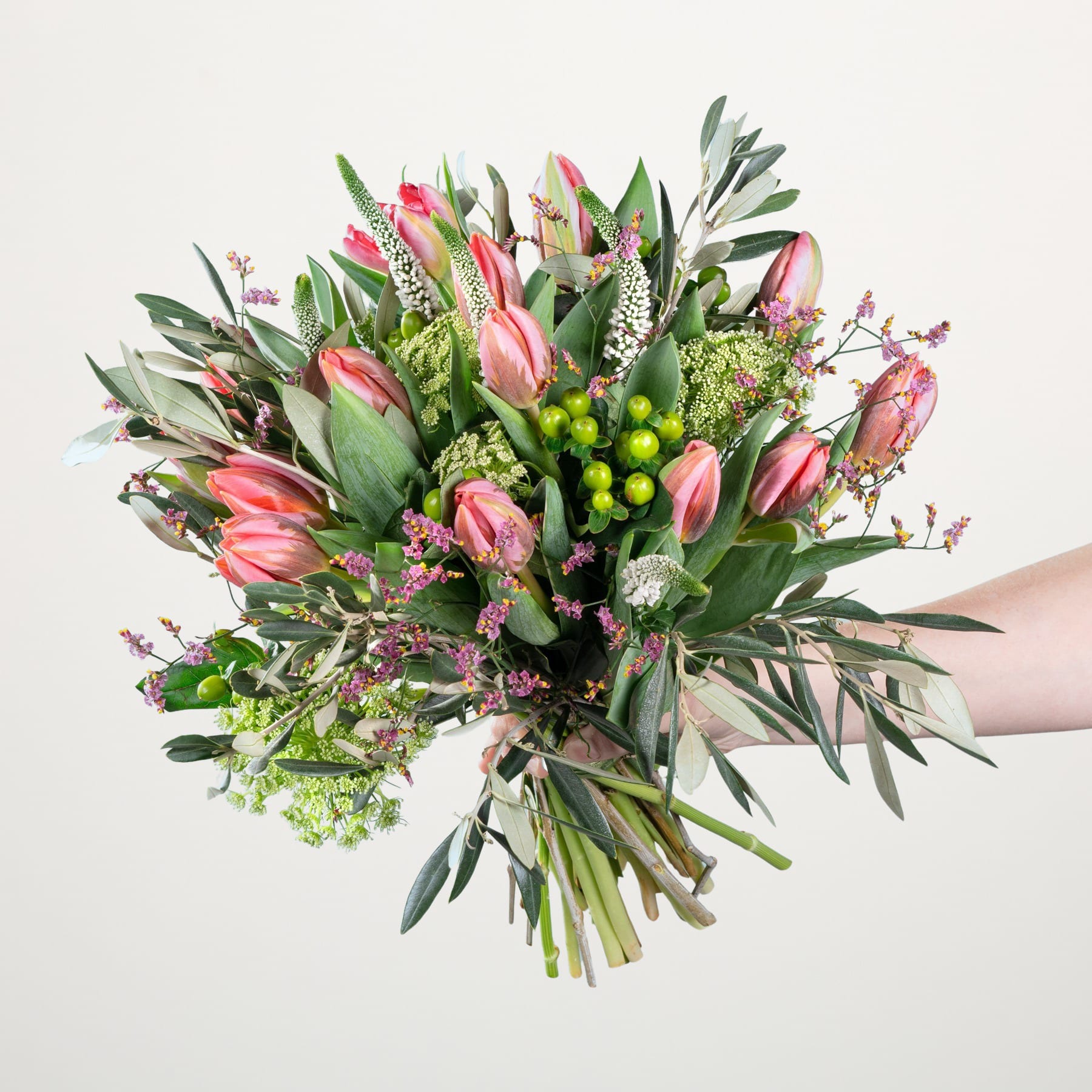 Ein leuchtender Strauß aus rosa Tulpen, grünem Laub und anderen bunten Blumen, arrangiert in einem Handdisplay vor einem schlichten weißen Hintergrund.