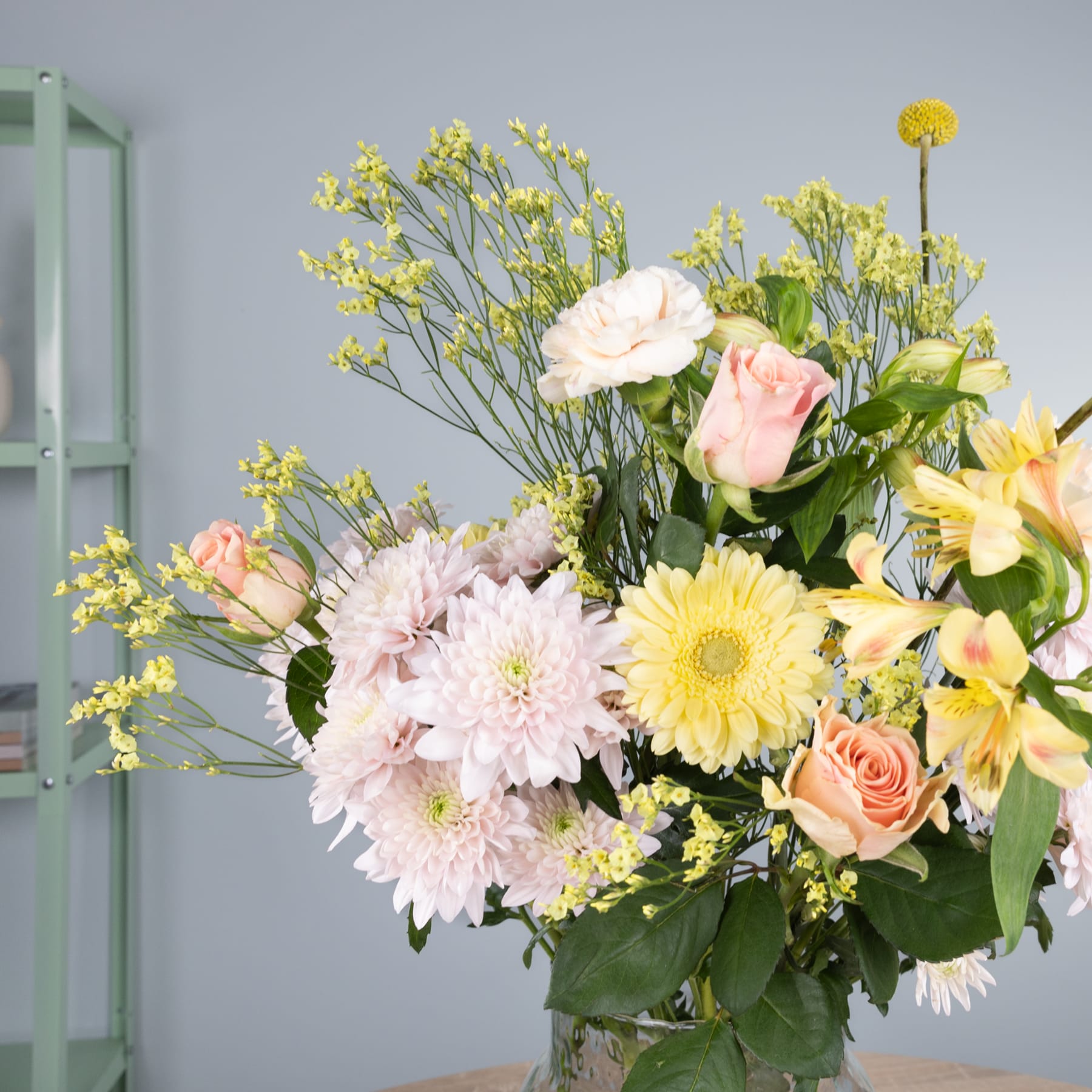 Ein farbenfroher Strauß verschiedener Blumen, darunter rosa und gelbe Gänseblümchen, Rosen und Grünpflanzen, steht im Vordergrund vor einem schlichten grauen Hintergrund.