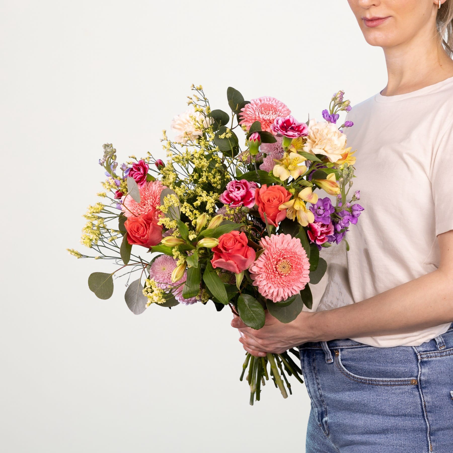 Ein leuchtender Strauß bunter Blumen, darunter rosa Nelken, gelbe Narzissen und violette Schwertlilien, wird von einer Person gehalten, die ein weißes Hemd und Jeans vor einem schlichten weißen Hintergrund trägt.
