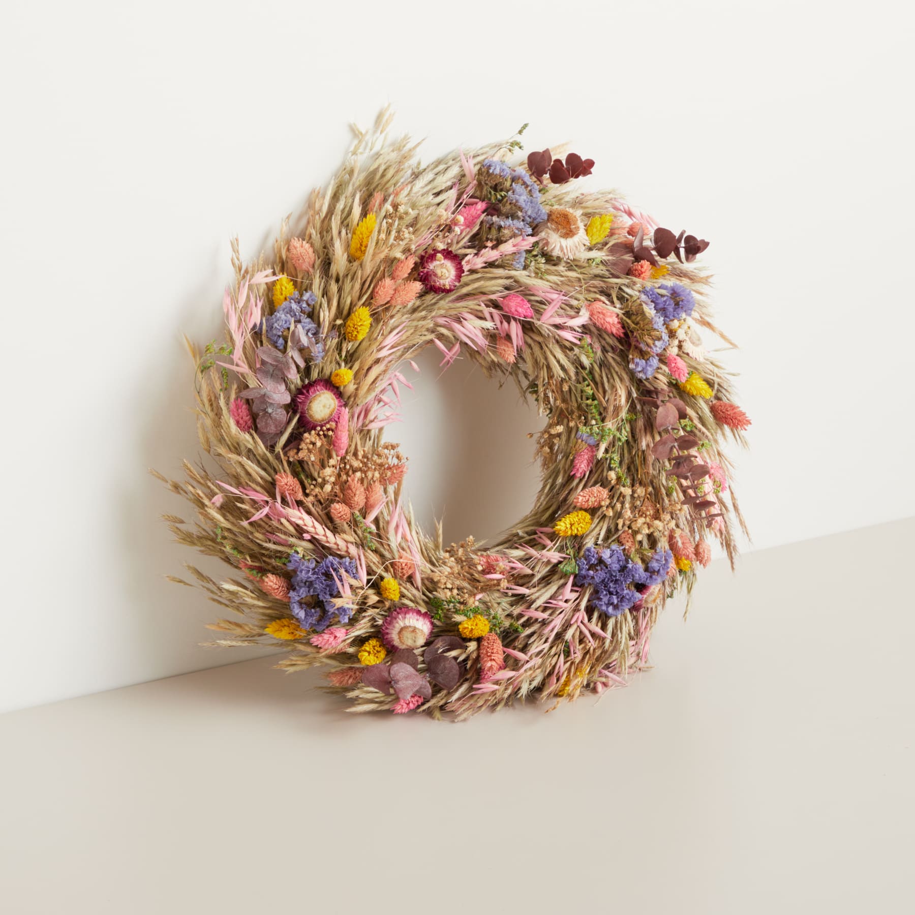 Ein lebendiger, runder Kranz aus getrockneten Blumen, Gräsern und anderen natürlichen Elementen wird vor einem schlichten, hellen Hintergrund präsentiert.