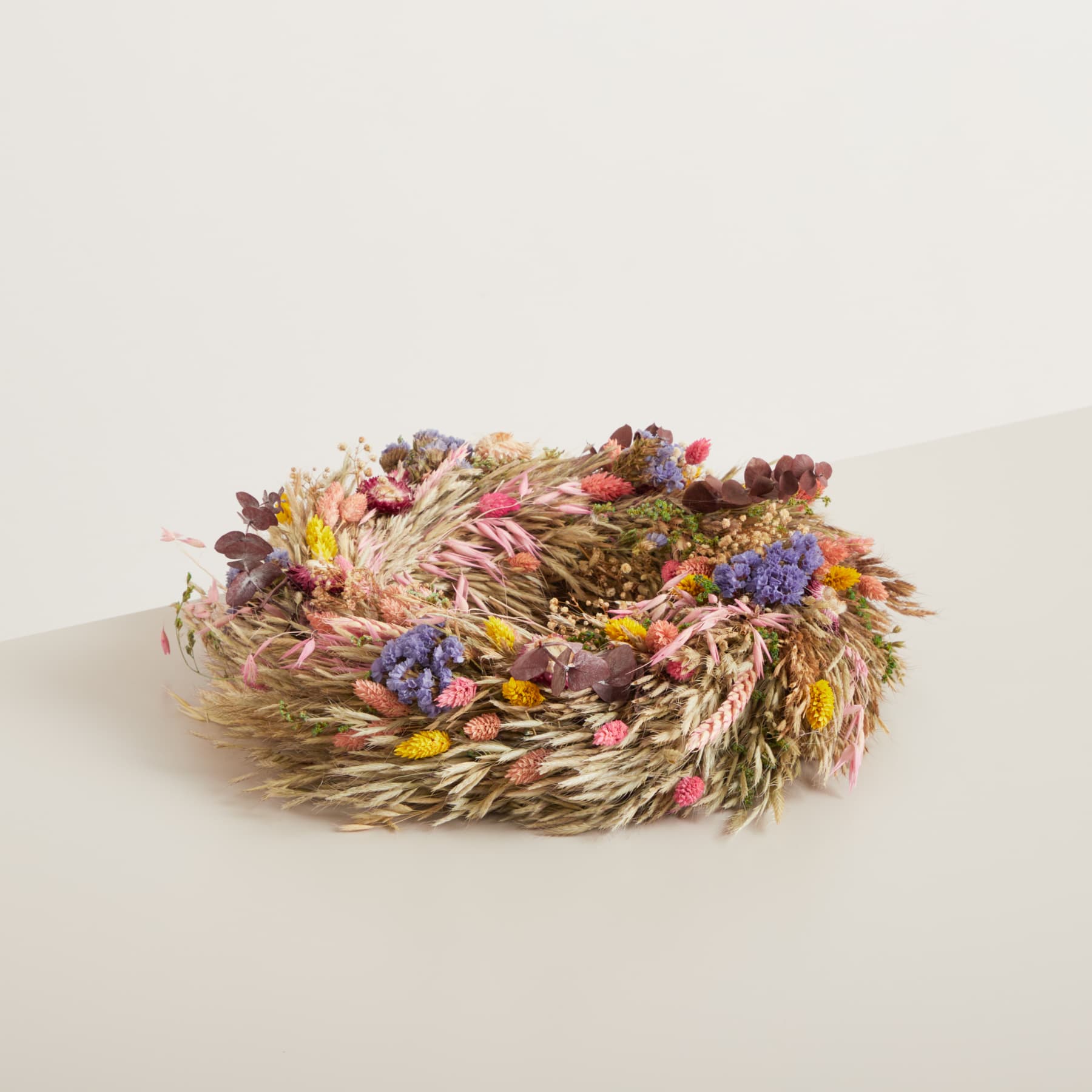 Auf einem schlichten weißen Hintergrund befindet sich ein bunter Kranz aus getrockneten Blumen, Gräsern und anderen Naturelementen.