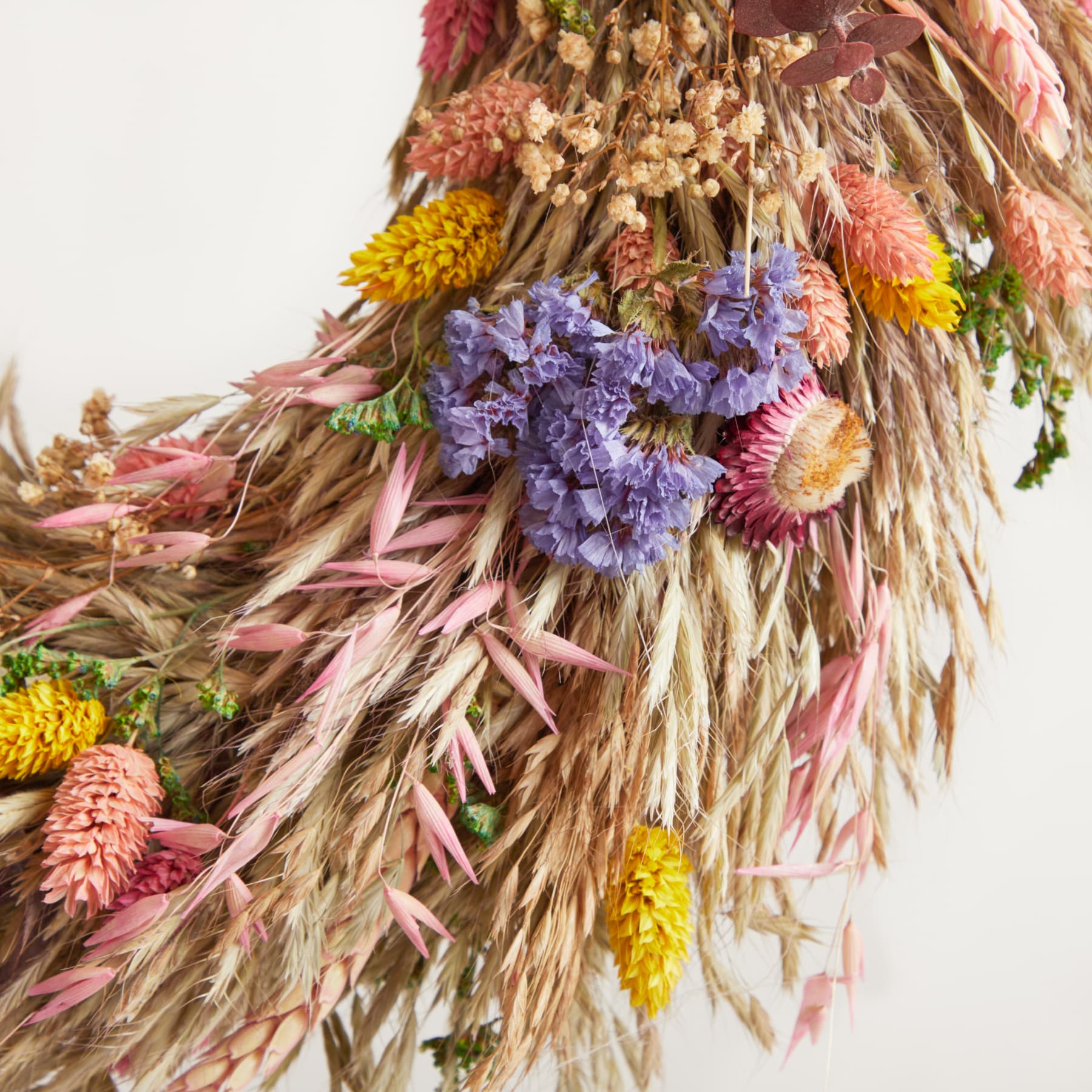 Ein farbenfroher Strauß getrockneter Blumen, darunter rosa und gelbe Blüten, verflochten mit Weizenhalmen, wodurch ein üppiges und strukturiertes Arrangement vor einem schlichten Hintergrund entsteht.