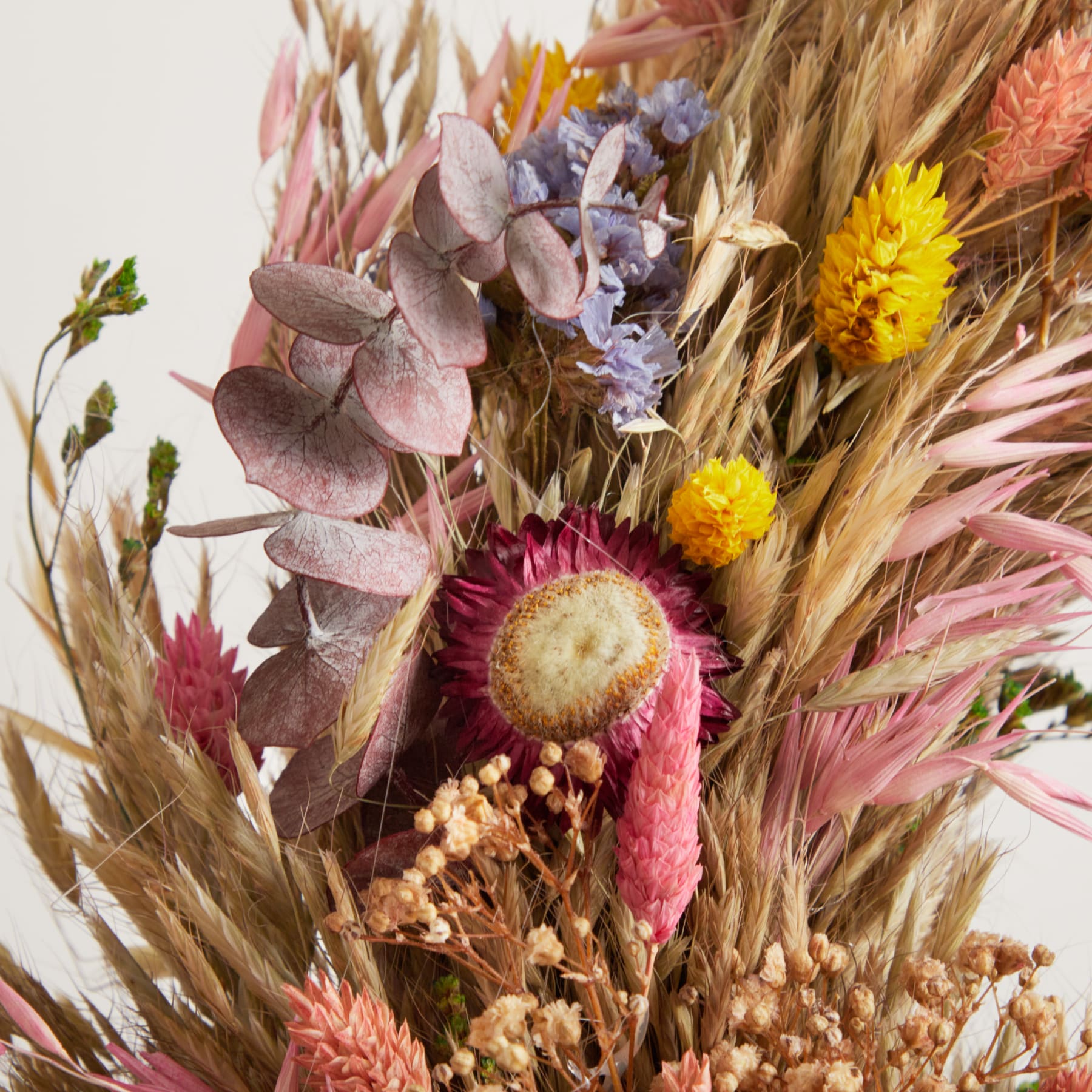 Ein farbenfroher Strauß getrockneter Blumen, darunter violette und gelbe Blüten, Weizenhalme und andere natürliche Elemente, vor einem schlichten Hintergrund.
