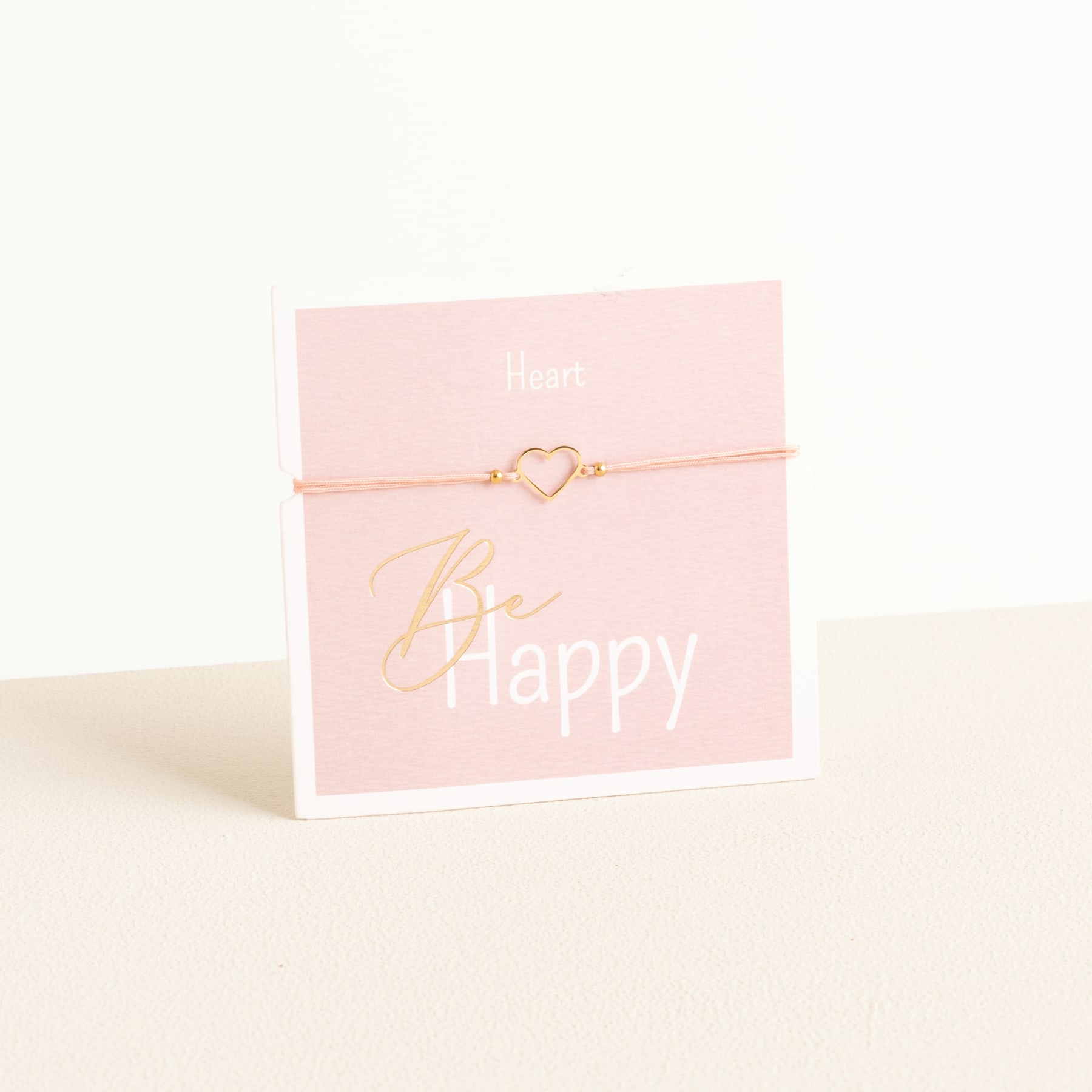 Auf einem hellen Untergrund liegt eine rosa Karte mit der Aufschrift „Be happy“ und einem herzförmigen Anhänger an einer zarten Schnur.