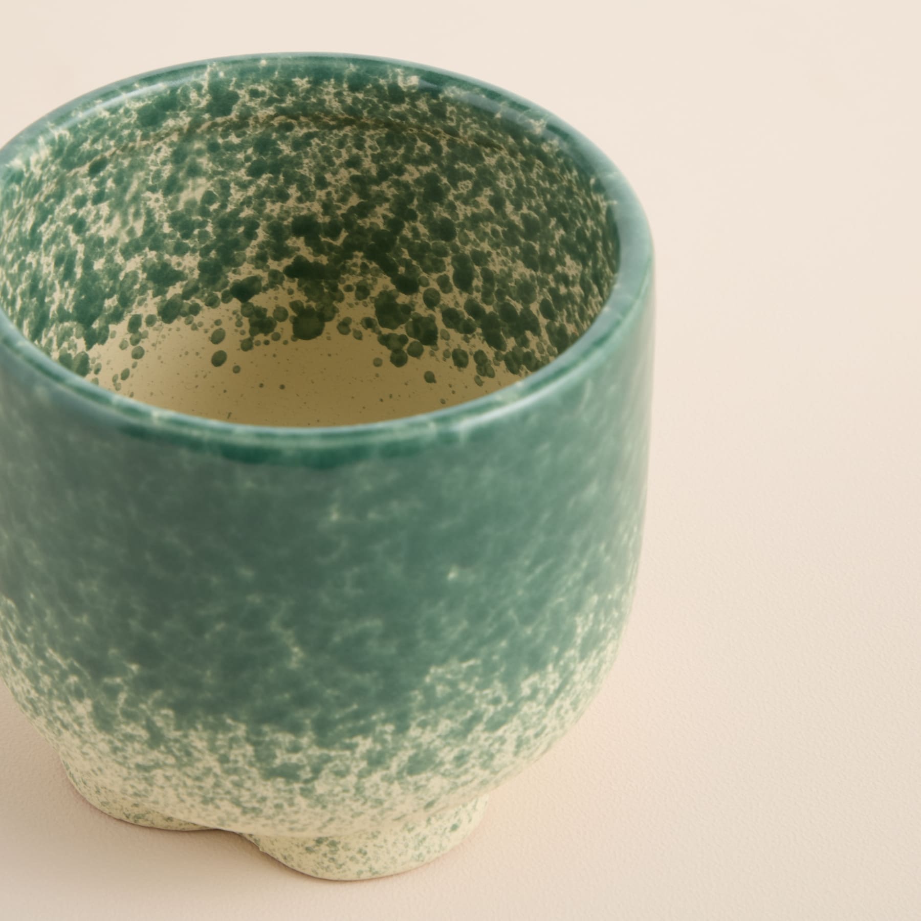Eine grün-beige Keramiktasse oder ein Keramikbecher mit gesprenkelter, strukturierter Oberfläche steht vor einem schlichten, hellen Hintergrund.