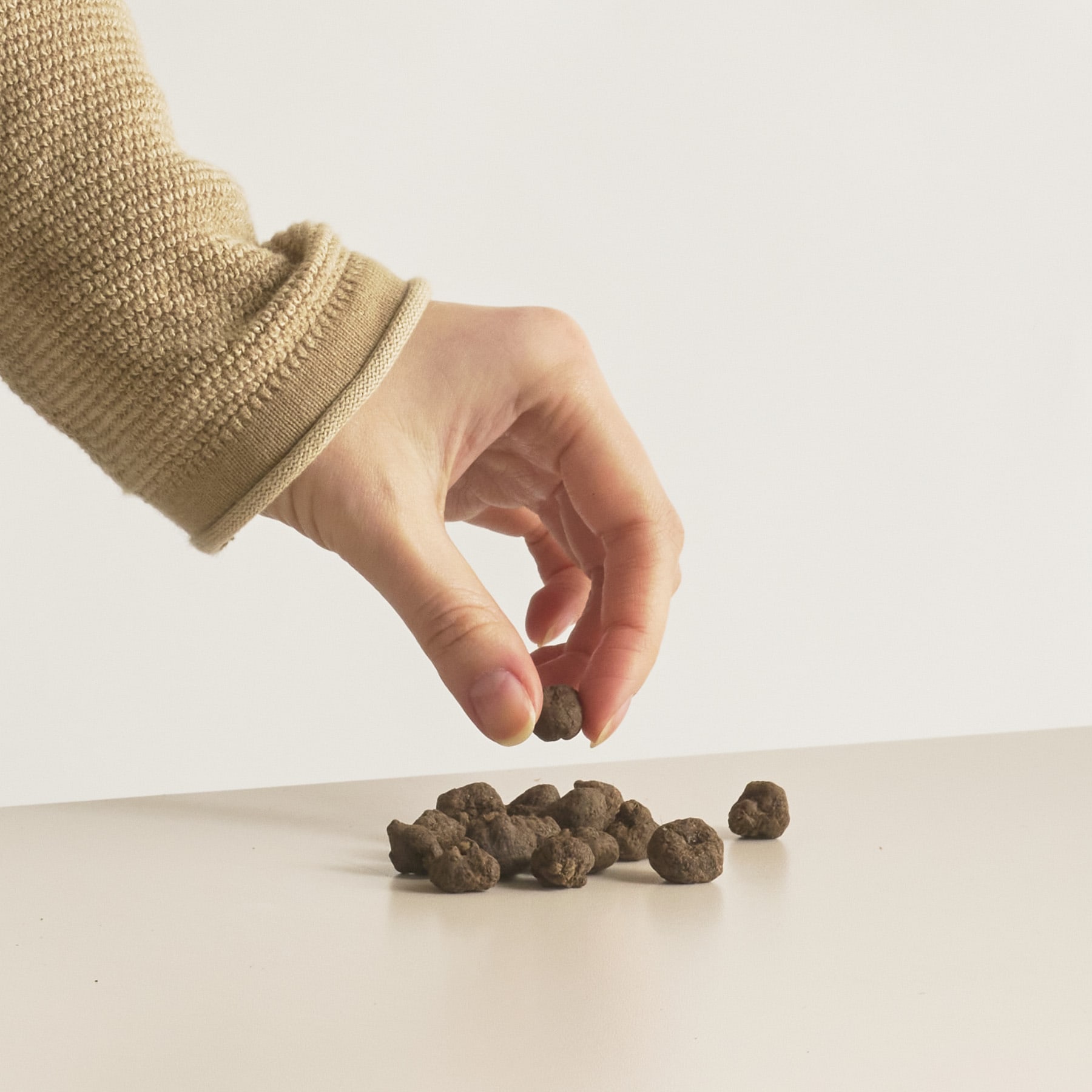 Eine Hand in einem beigen Pullover streckt sich nach unten und berührt einen Haufen kleiner, unregelmäßig geformter Steine auf einer schlichten weißen Oberfläche.