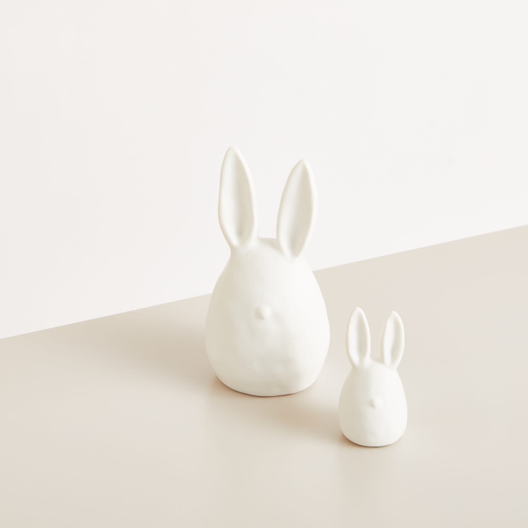 Zwei weiße Keramik-Kaninchenfiguren unterschiedlicher Größe sitzen auf einer schlichten, hellen Oberfläche vor einem neutralen Hintergrund.