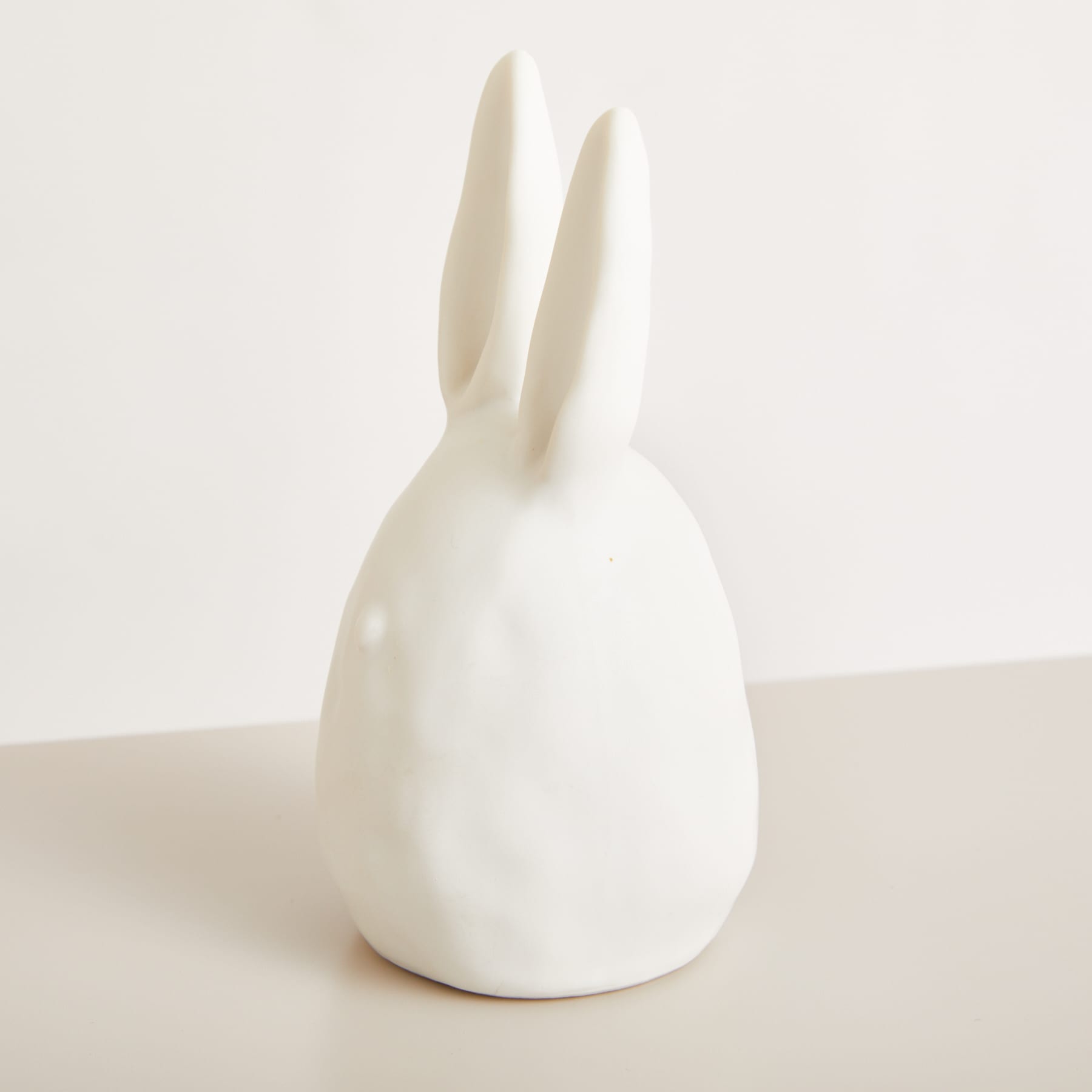Eine einfache, minimalistische weiße Kaninchenfigur aus Keramik sitzt auf einer hellen Oberfläche vor einem schlichten Hintergrund.