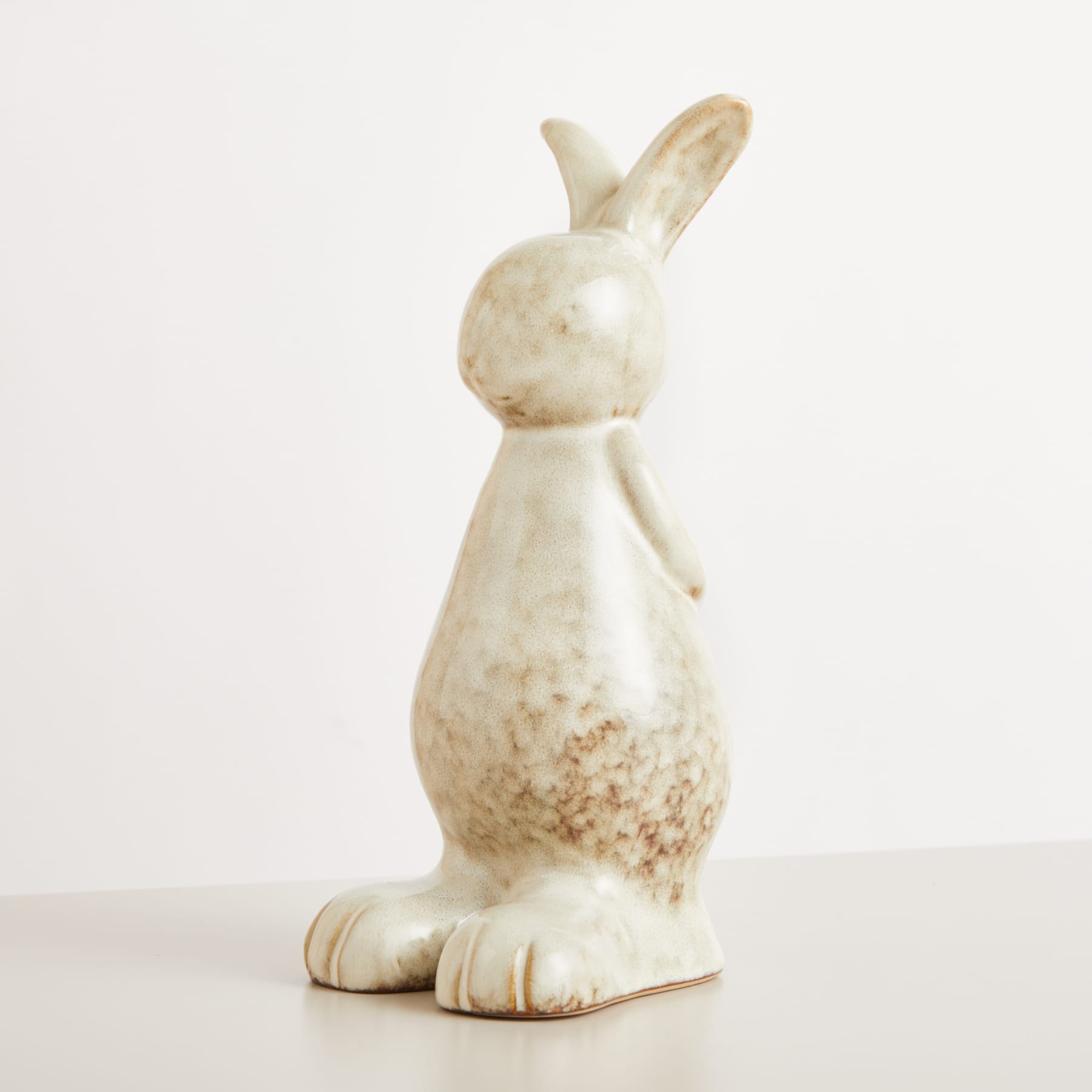 Eine Keramikfigur eines Kaninchens mit einer gesprenkelten beige-weißen Glasur steht allein auf einem schlichten weißen Hintergrund.