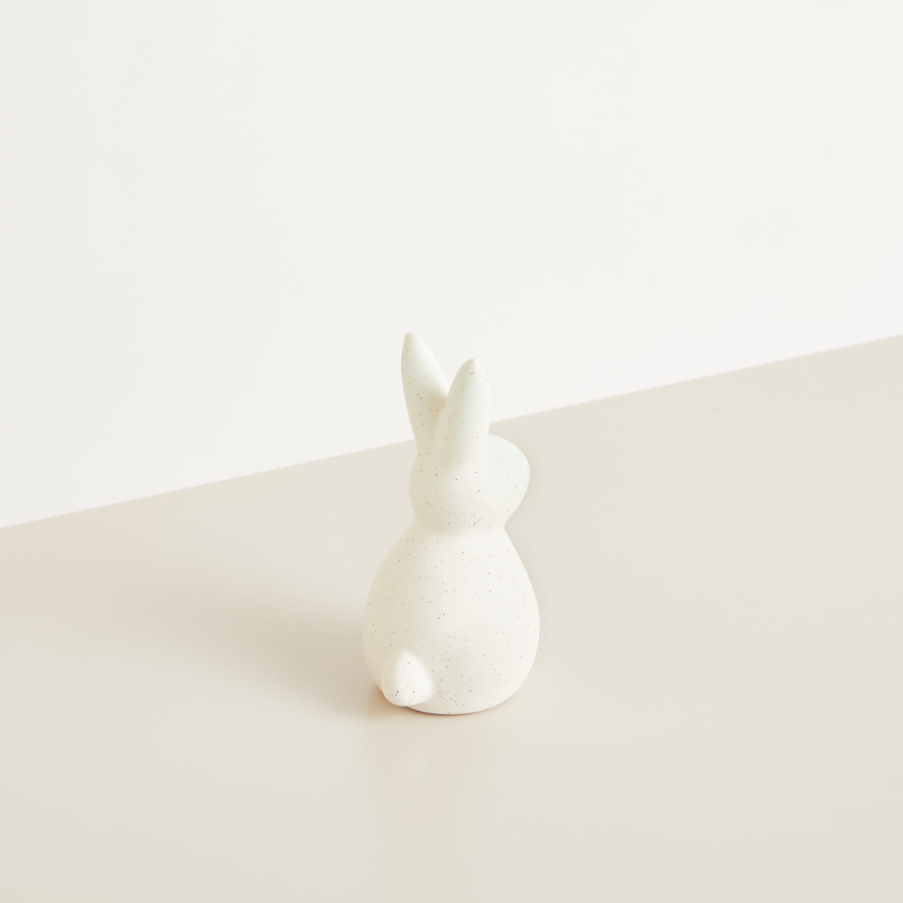 Eine weiße Kaninchenfigur aus Keramik sitzt auf einer schlichten, hellen Oberfläche vor einem neutralen Hintergrund.