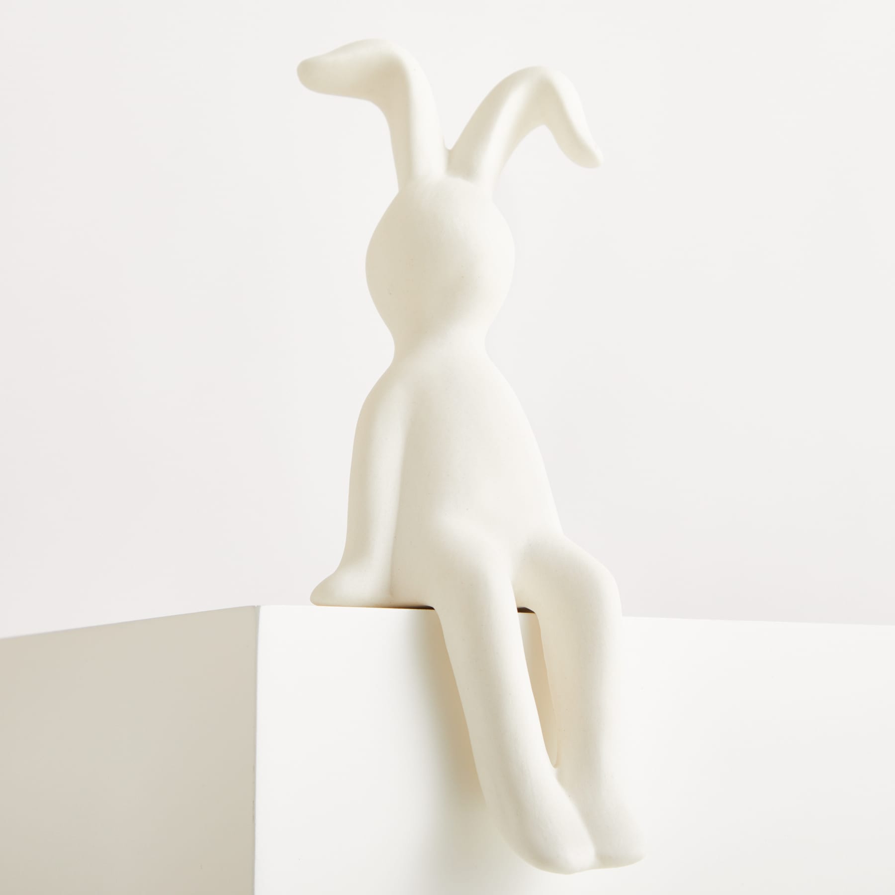 Eine weiße, minimalistische Skulptur einer kaninchenähnlichen Figur, die auf einer weißen Plattform vor einem schlichten weißen Hintergrund sitzt.