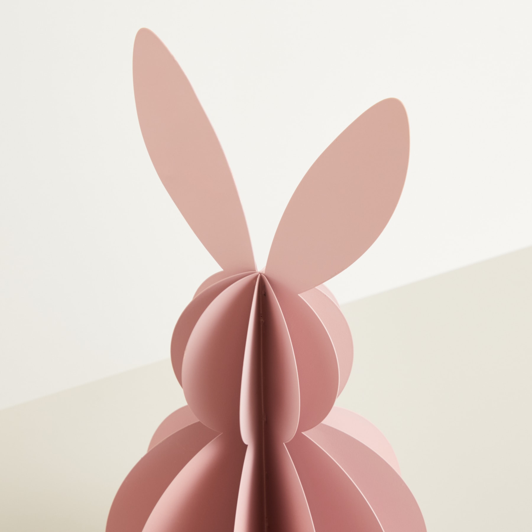 Vor einem schlichten, hellen Hintergrund steht eine rosa Papierskulptur, die einem Hasenkopf mit großen Ohren ähnelt.