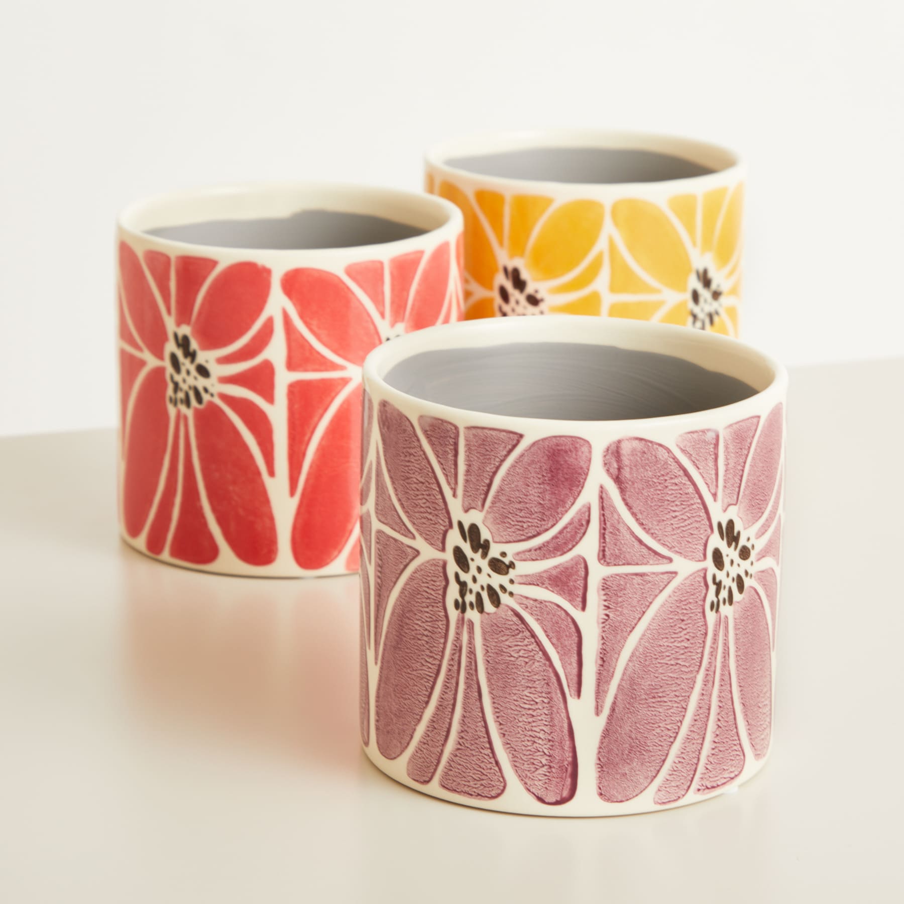 Drei Keramikbecher oder -tassen mit leuchtenden Blumenmustern in Rot-, Orange- und Rosatönen auf schlichtem weißem Hintergrund.