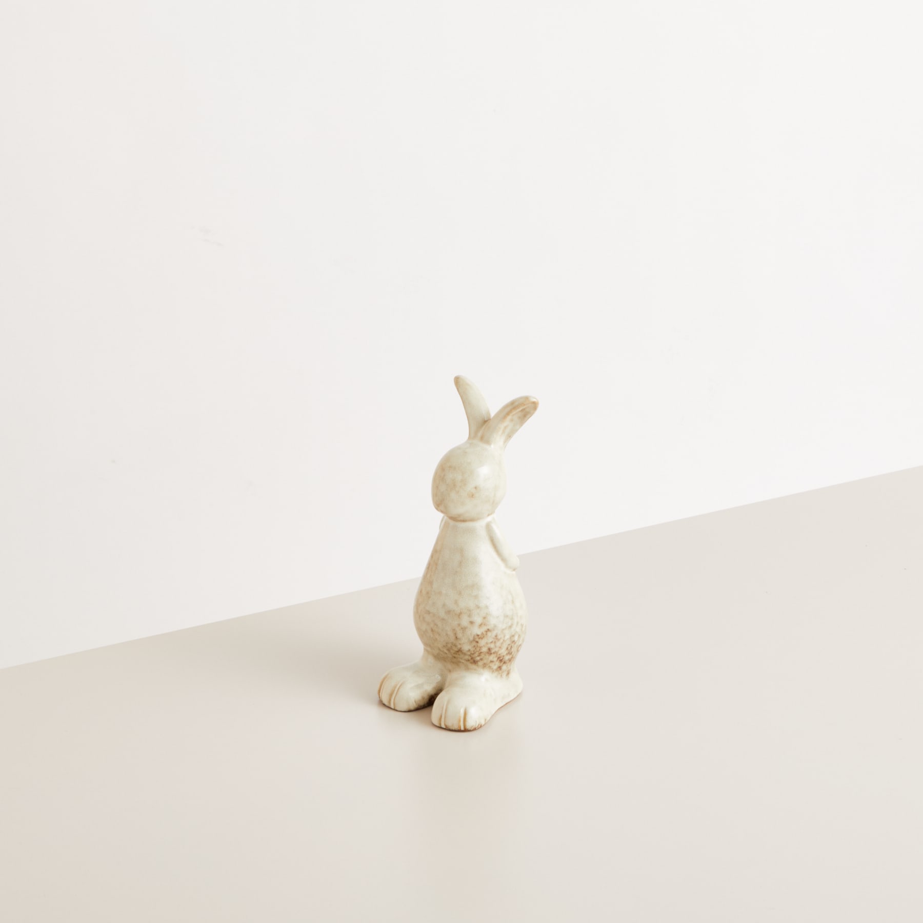 Eine flauschige weiße Kaninchenfigur sitzt auf einer schlichten, hellen Oberfläche vor einem neutralen Hintergrund.