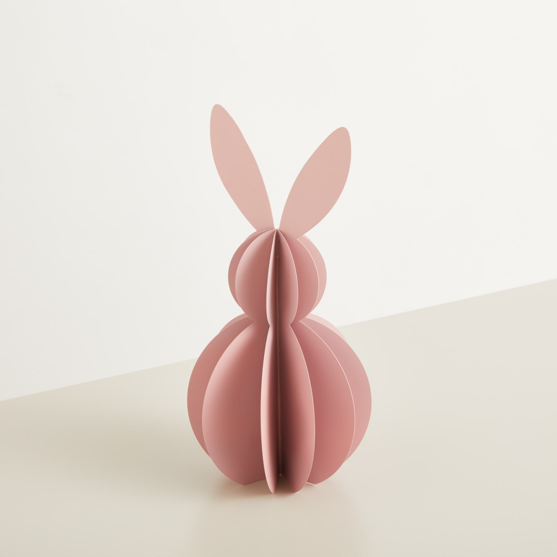 Eine rosafarbene, papierartige Skulptur mit hasenartigen Ohren steht auf einer hellen Oberfläche vor einem schlichten Hintergrund.
