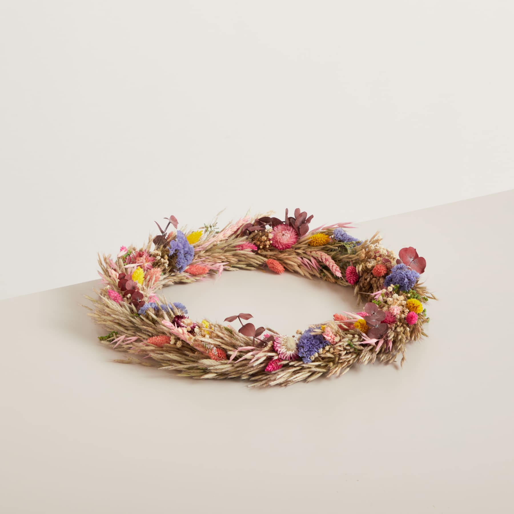 Auf einer schlichten, hellen Oberfläche liegt ein bunter Kranz aus getrockneten Blumen, Blättern und anderen Naturelementen.