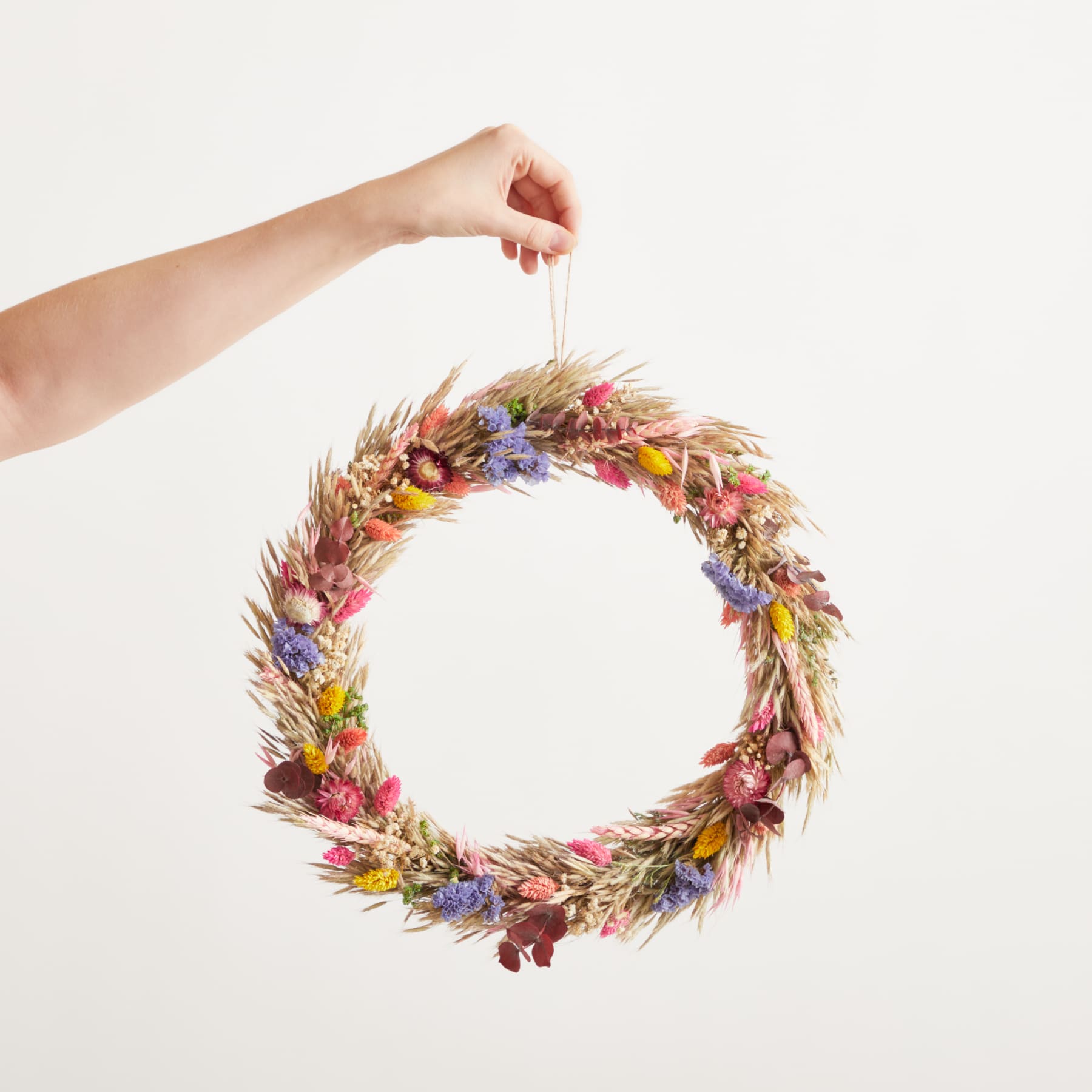 Eine Hand hält einen Kranz aus getrockneten Blumen und Blättern vor einem schlichten weißen Hintergrund.