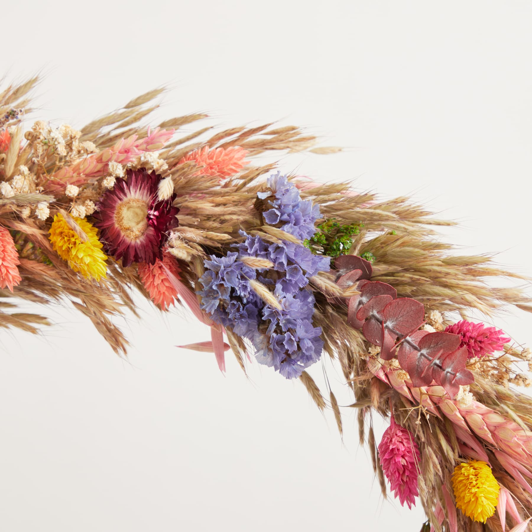 Ein farbenfroher Strauß getrockneter Blumen, darunter rosa und violette Blüten, Weizenhalme und andere natürliche Elemente, arrangiert vor einem schlichten weißen Hintergrund.