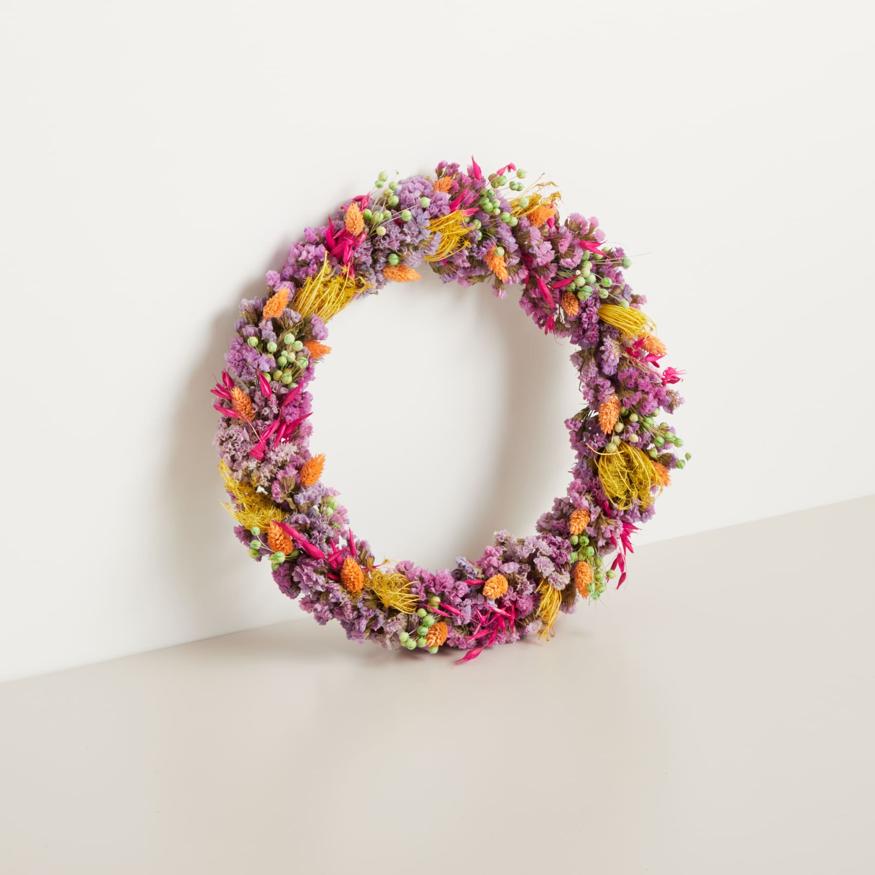 Ein leuchtender, runder Kranz aus verschiedenen bunten Blumen, darunter rosa, gelbe und violette Blüten, wird vor einem schlichten weißen Hintergrund präsentiert.