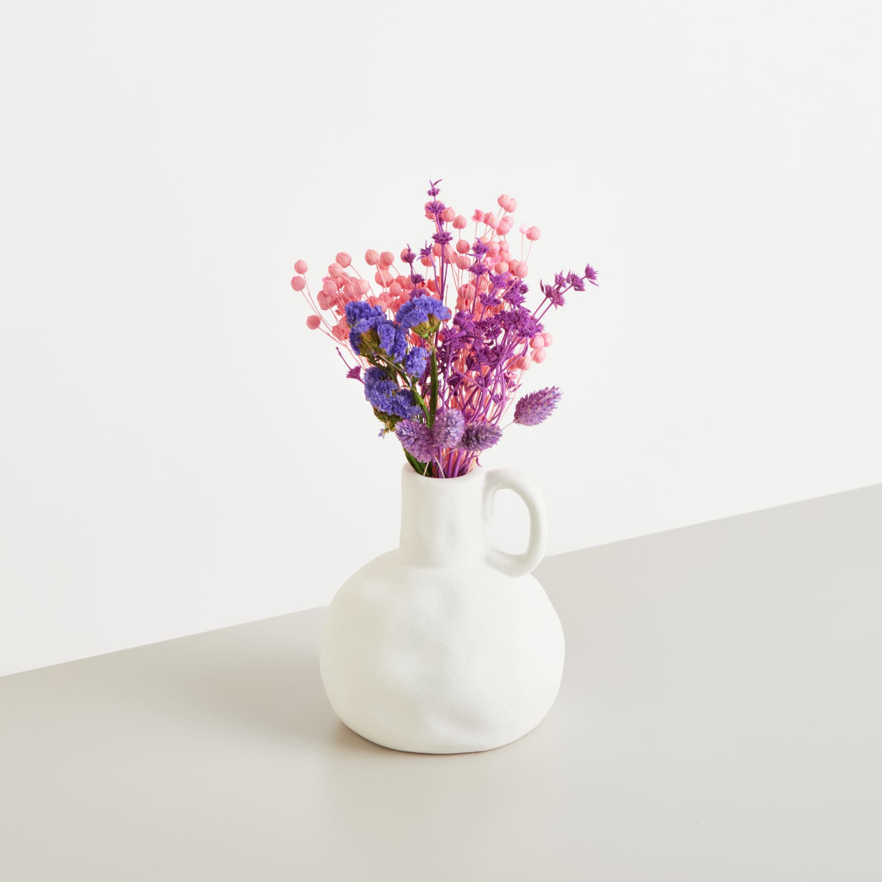 Eine weiße Keramikvase mit einem farbenfrohen Strauß getrockneter Blumen in Lila und Rosa steht auf einer schlichten weißen Fläche vor einem schlichten weißen Hintergrund.