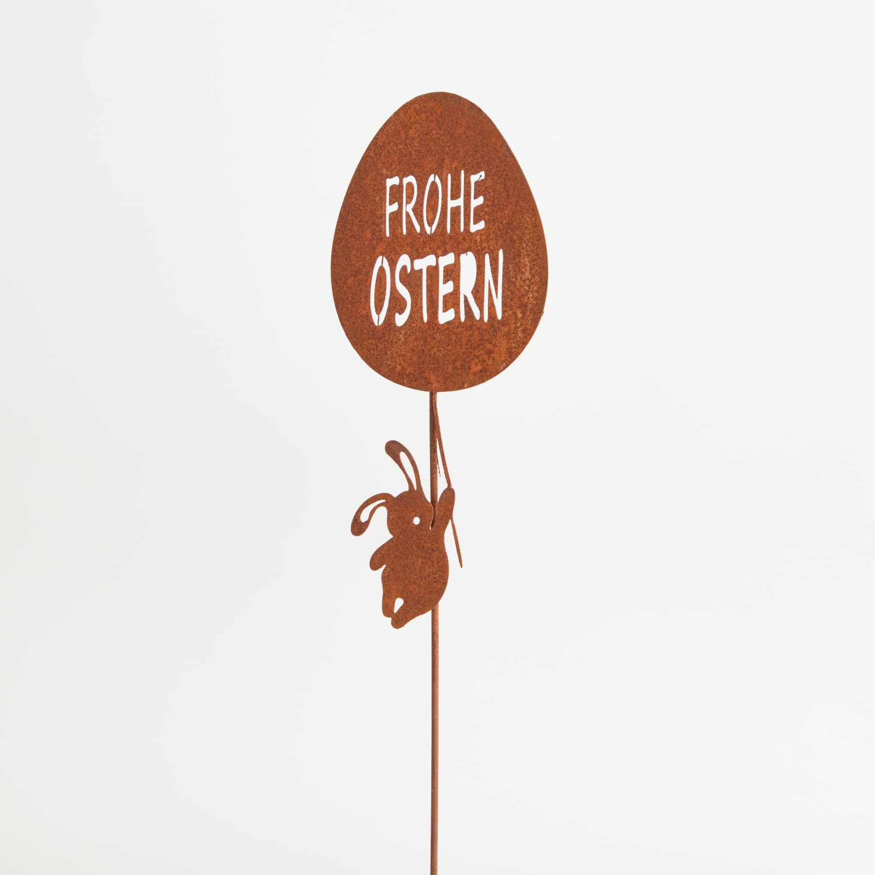 Ein großes, rostfarbenes Schild in Form eines Osterei mit dem deutschen Text „FROHE OSTERN“ wird von einer kleinen, skurrilen Hasenfigur hochgehalten.