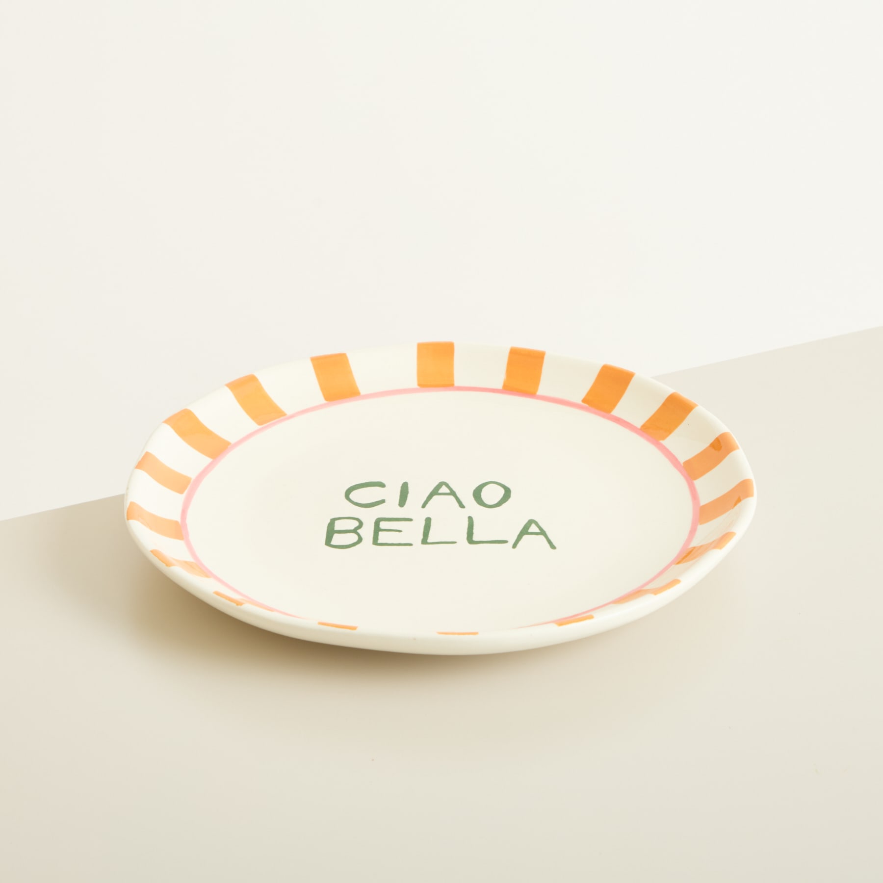 Ein weißer Keramikteller mit dem Text „CIAO BELLA“ und einem orange-gelb gestreiften Rand.