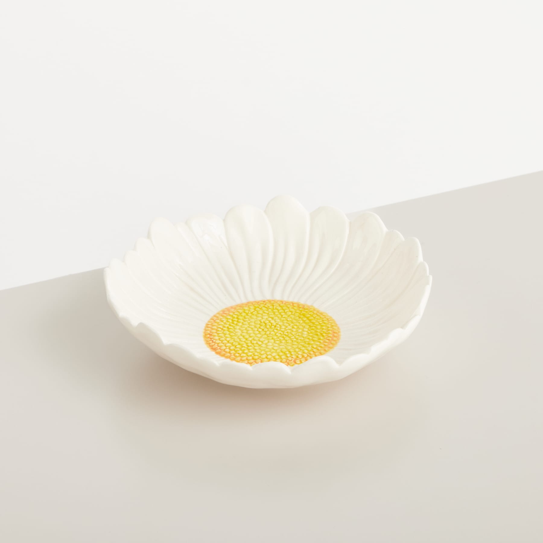 Eine weiße Keramikschale mit einer leuchtend gelben Mitte, die an eine Gänseblümchenblüte erinnert, steht auf einer schlichten, hellen Oberfläche.