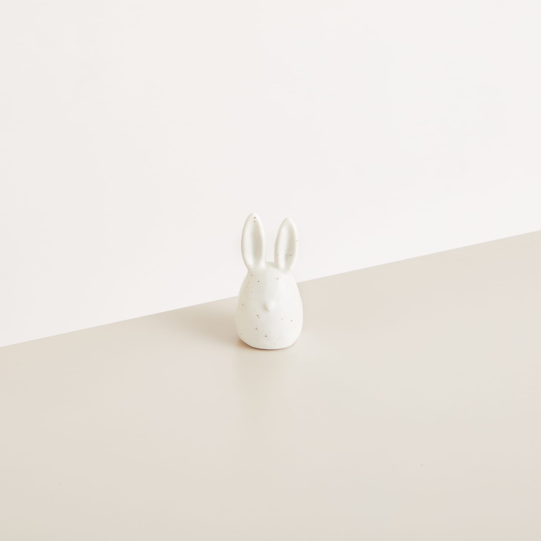 Eine kleine, weiße Kaninchenfigur aus Keramik sitzt auf einer schlichten, beigen Oberfläche vor einem hellen Hintergrund.