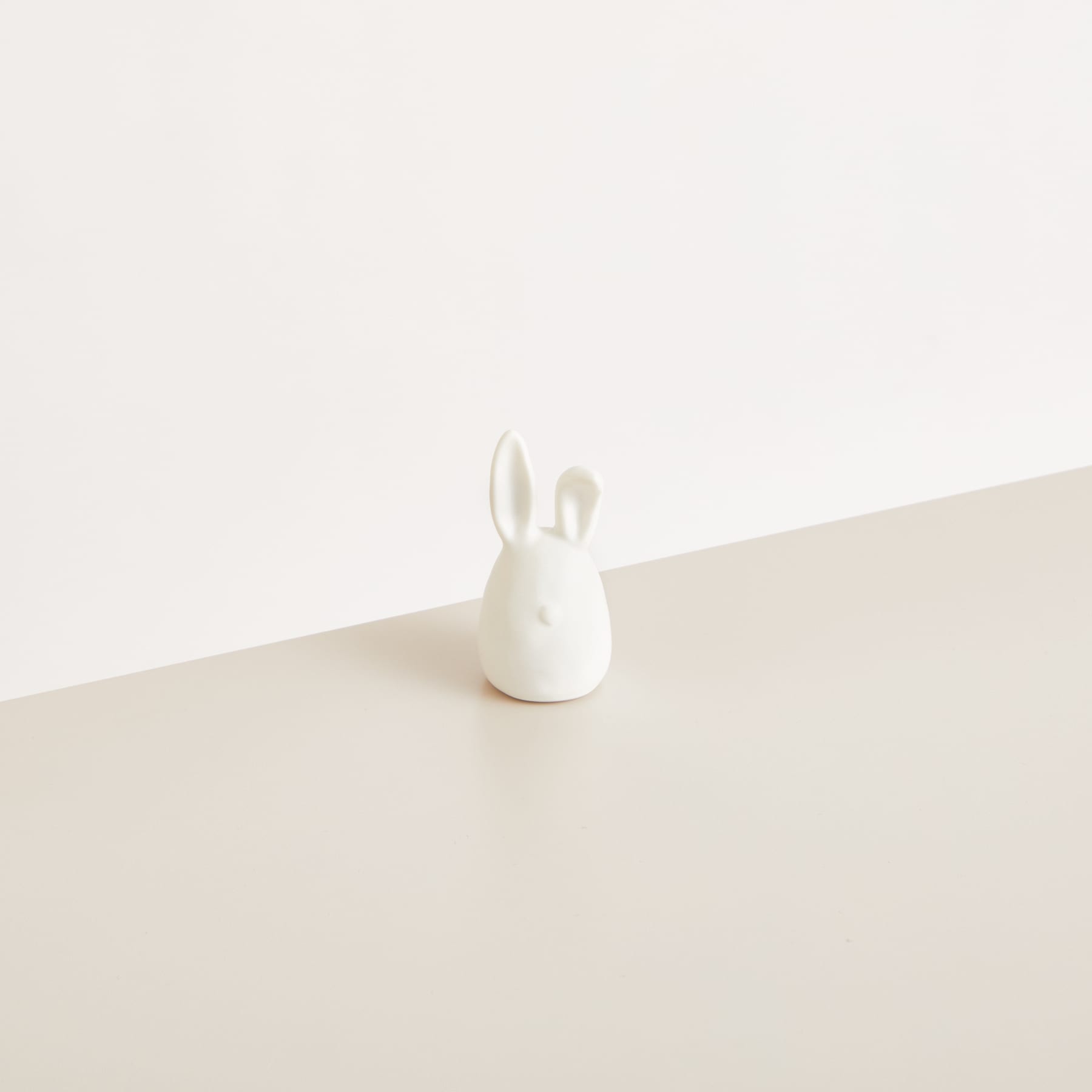 Eine einfache, minimalistische Kaninchenfigur aus Keramik sitzt auf einer schlichten, hellen Oberfläche vor einem neutralen Hintergrund.