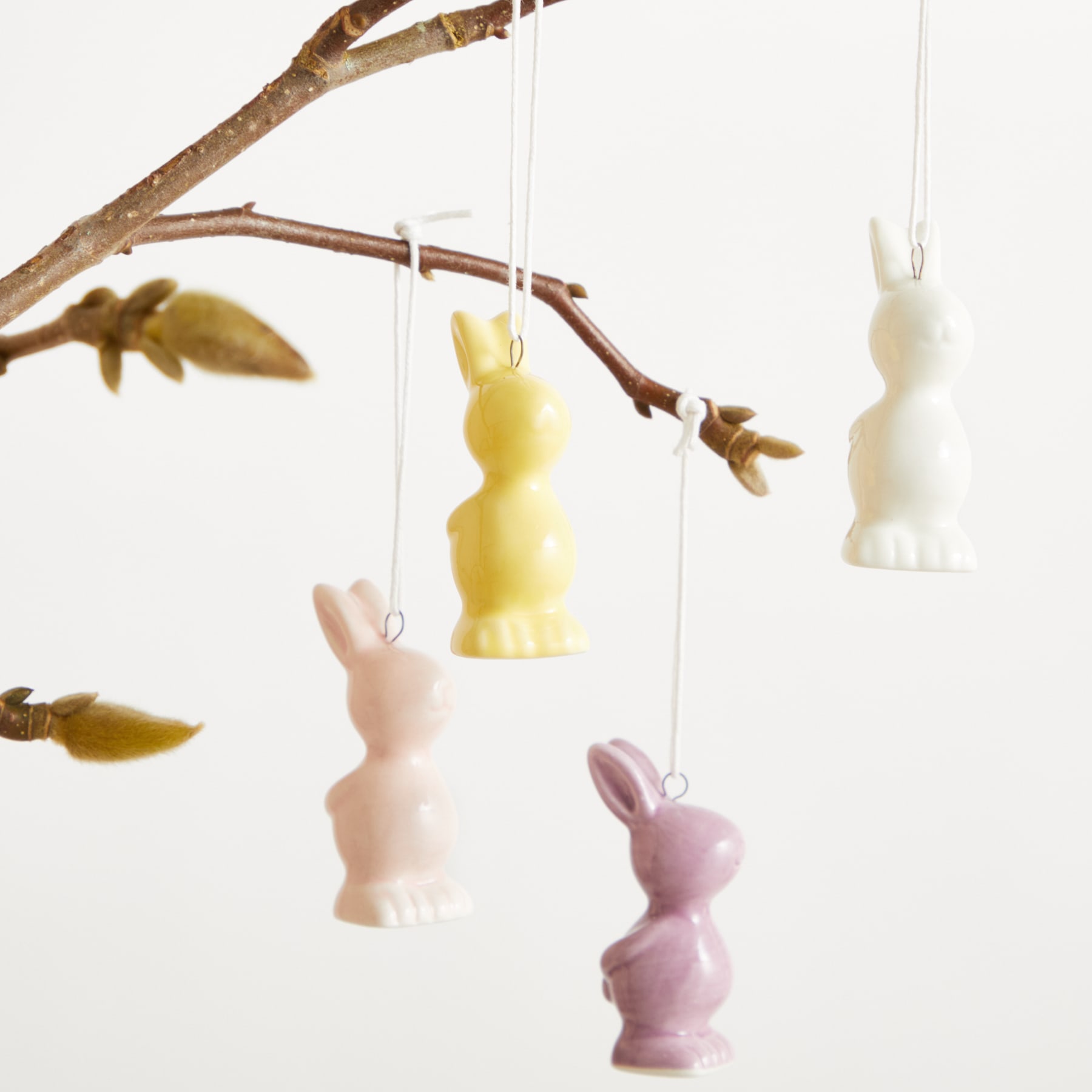 Das Bild zeigt ein Mobile oder eine hängende Dekoration mit mehreren kleinen, bunten Hasenfiguren aus Keramik oder Porzellan, die vor einem schlichten weißen Hintergrund an einem Ast hängen.